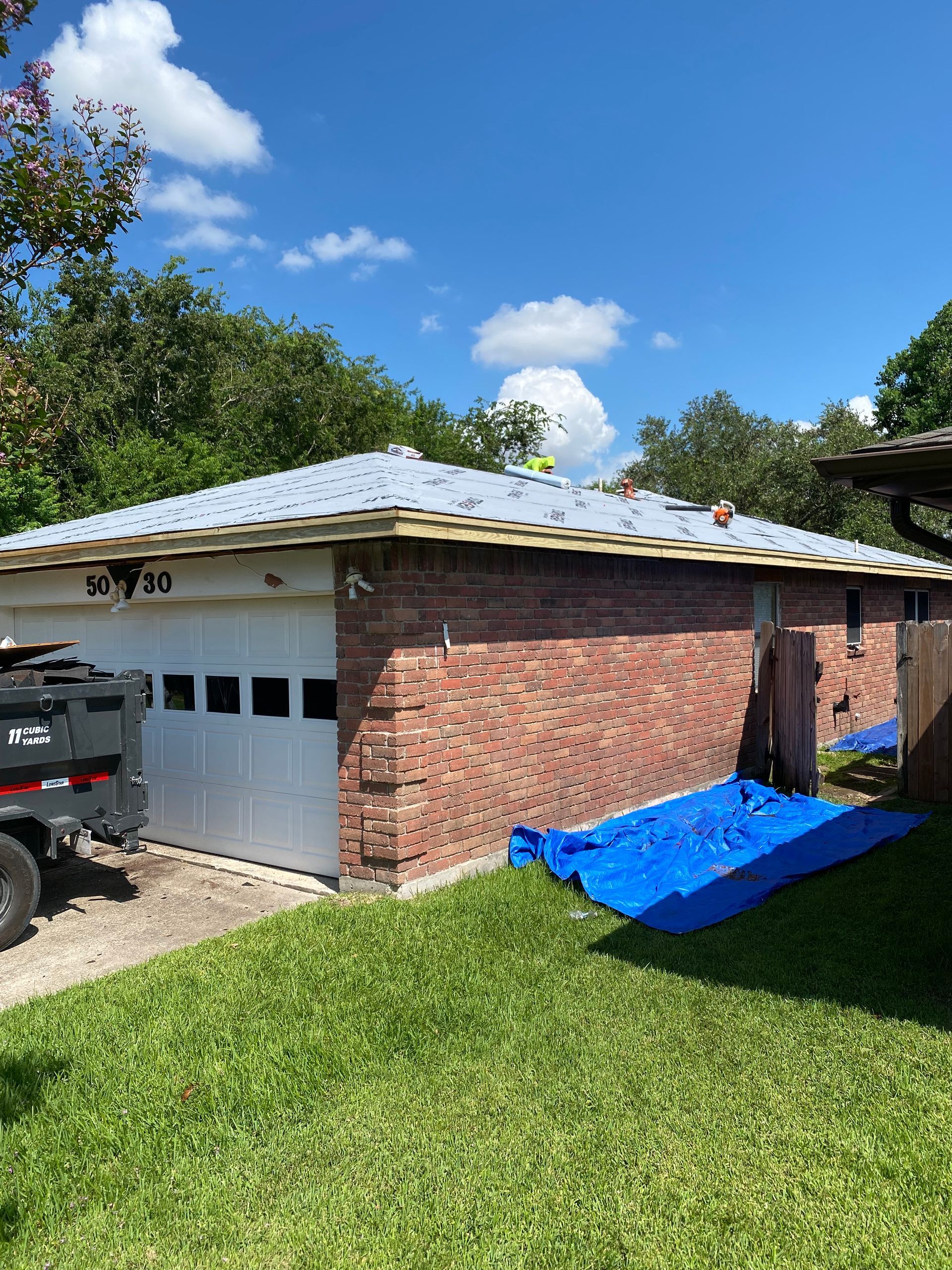 Garage with white door, brick walls, and roof being worked on, blue tarp on ground.