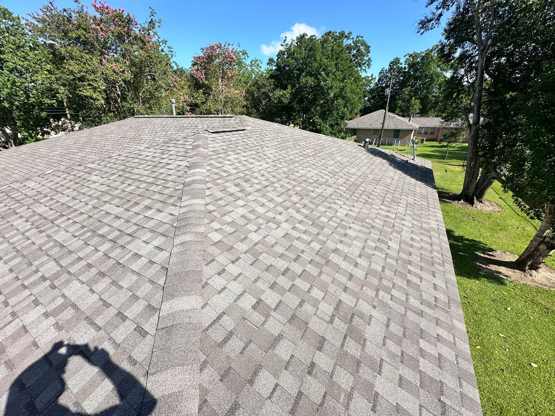 View of a gray shingled roof with trees in the background under a blue sky.