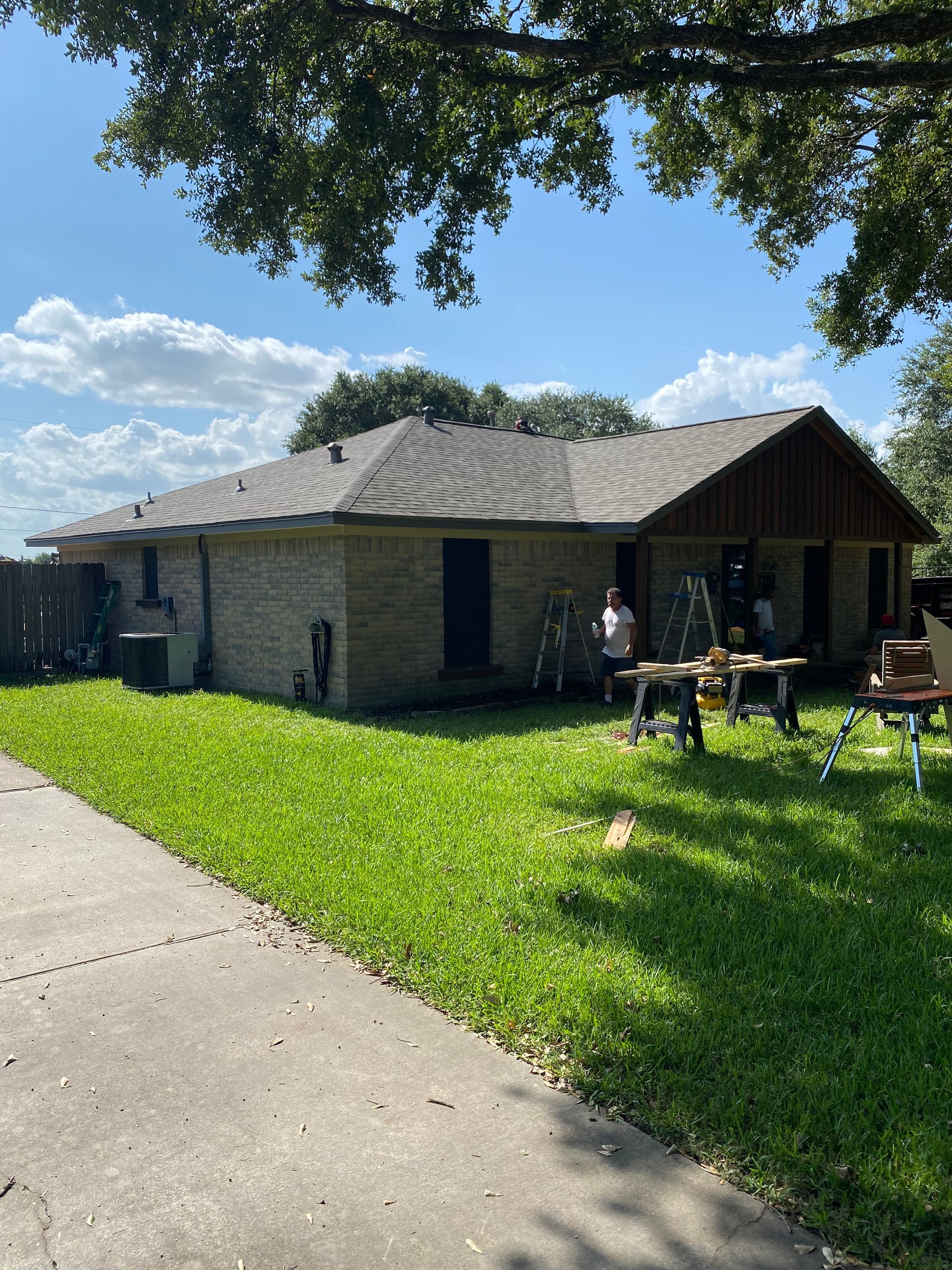 Brick house with dark roof and green lawn. People and tools outside. Sunny day.
