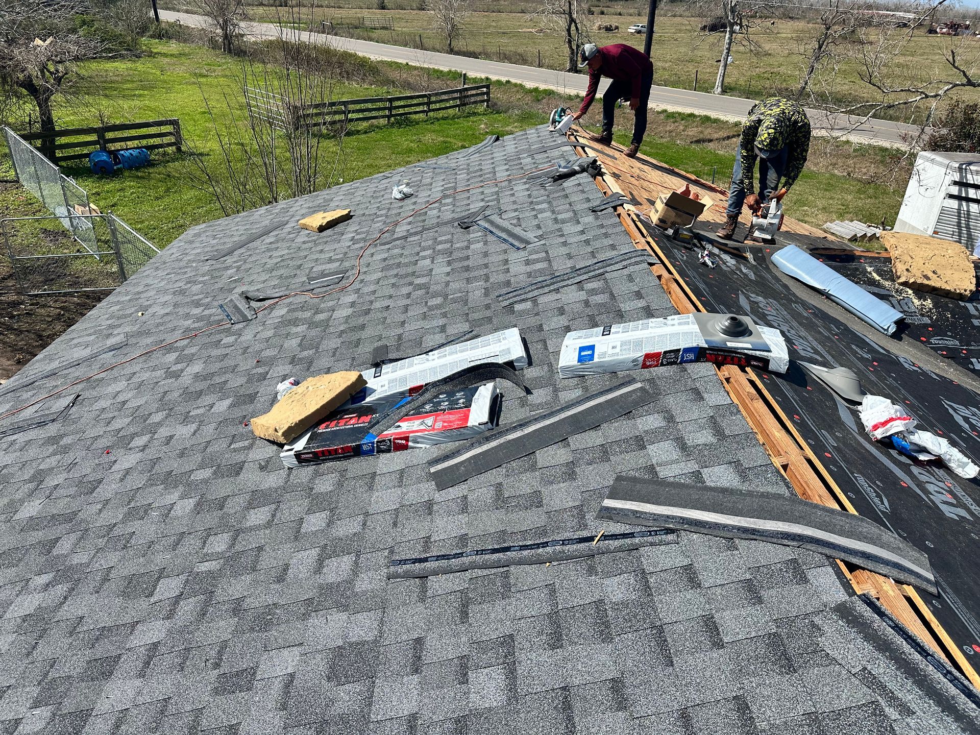 Two workers repairing a gray shingled roof, removing old shingles and installing new ones on a sunny day.