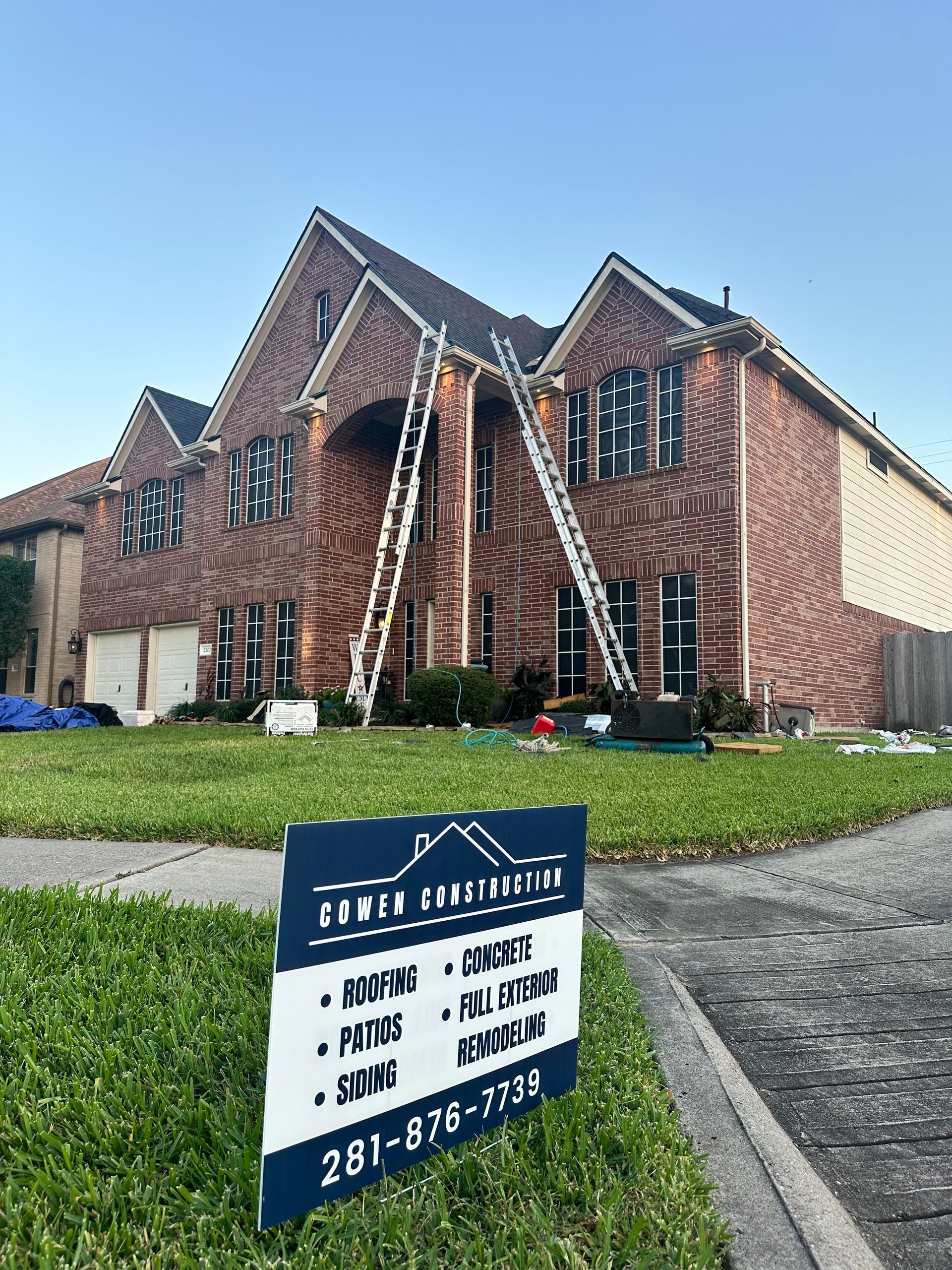Two-story brick house with ladders, construction sign, and roofing work in progress on a sunny day.