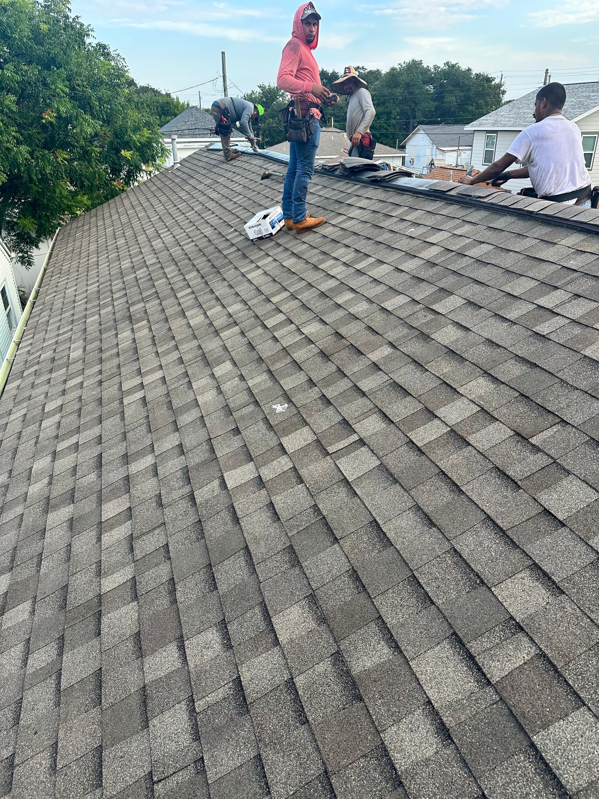 Roofers working on a shingled roof, trees and buildings in the background. Overcast sky.