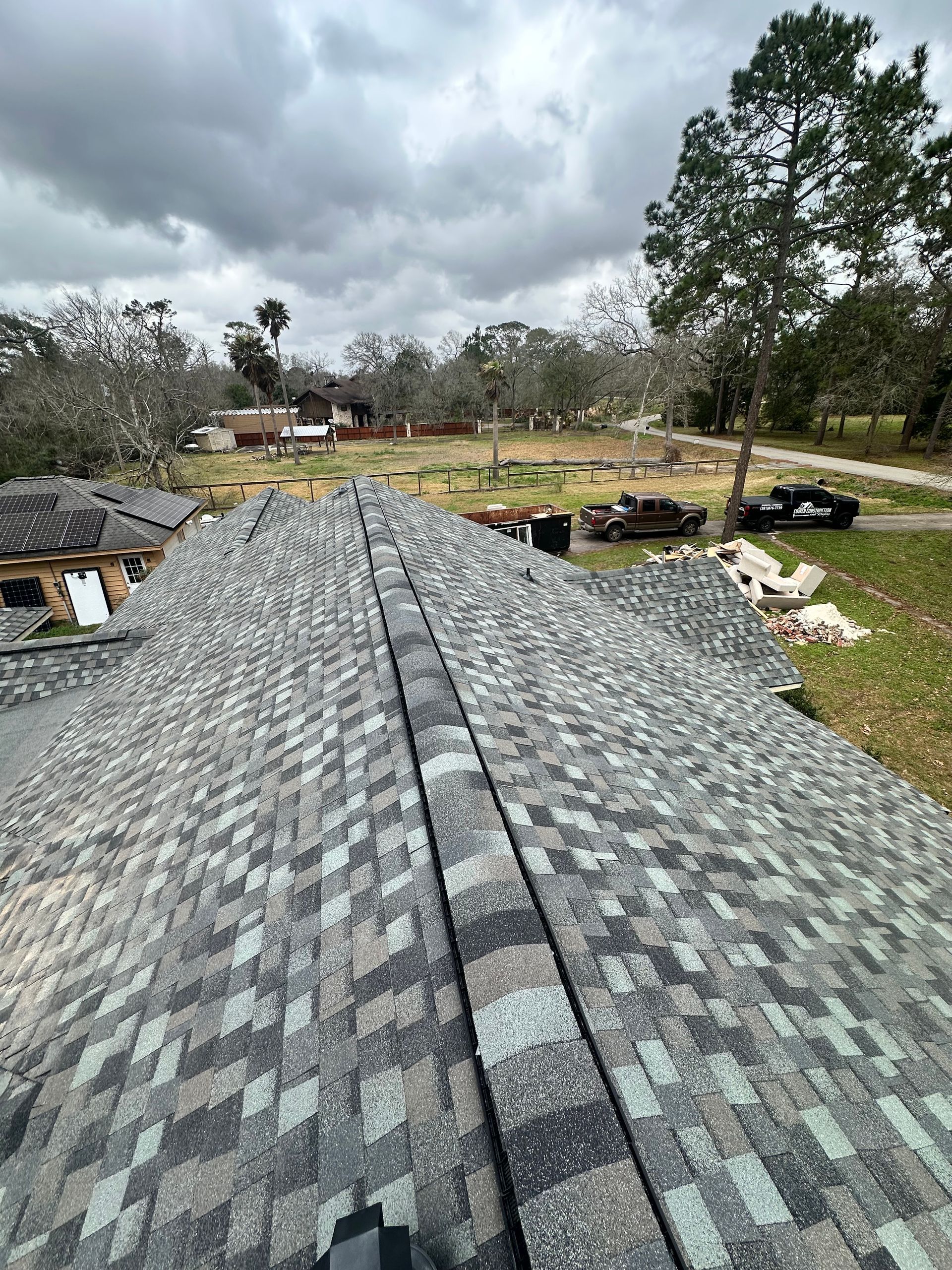 Overhead view of a newly shingled roof with dark gray shingles. Overcast day with trees and houses in the background.