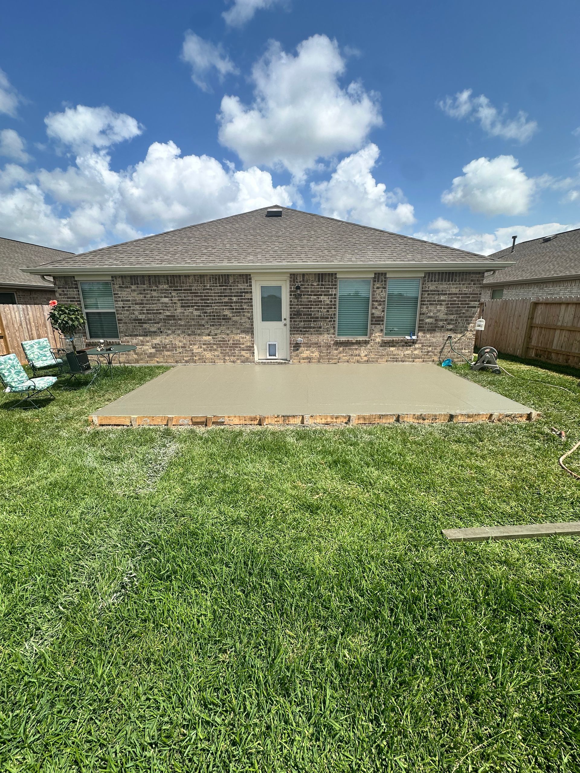 Backyard view of a brick house with concrete patio and green grass, blue sky with clouds.