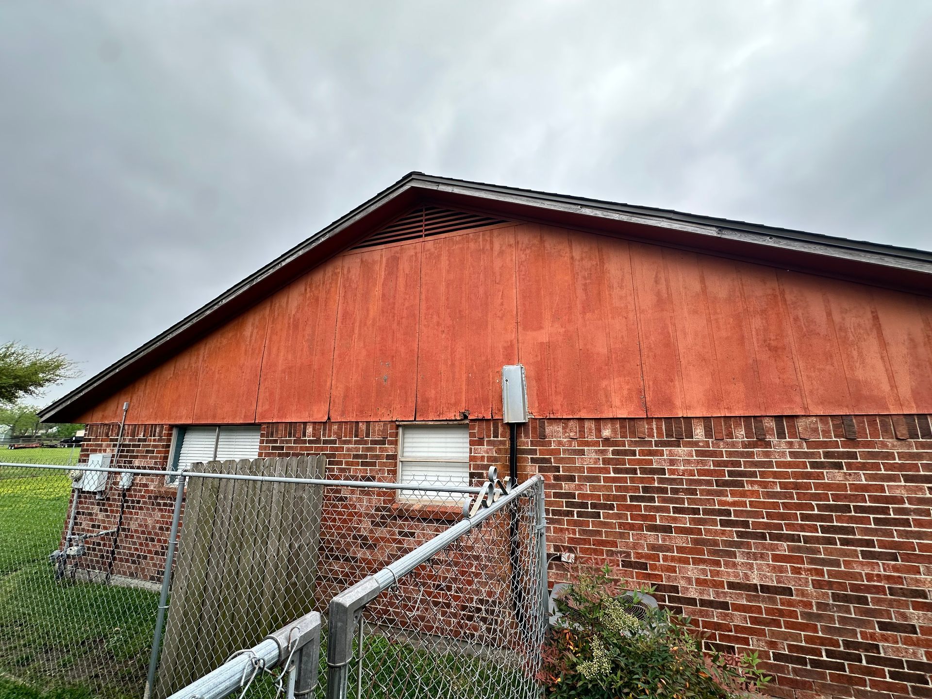 Brick building with a red wooden gable under an overcast sky, behind a chain-link fence.
