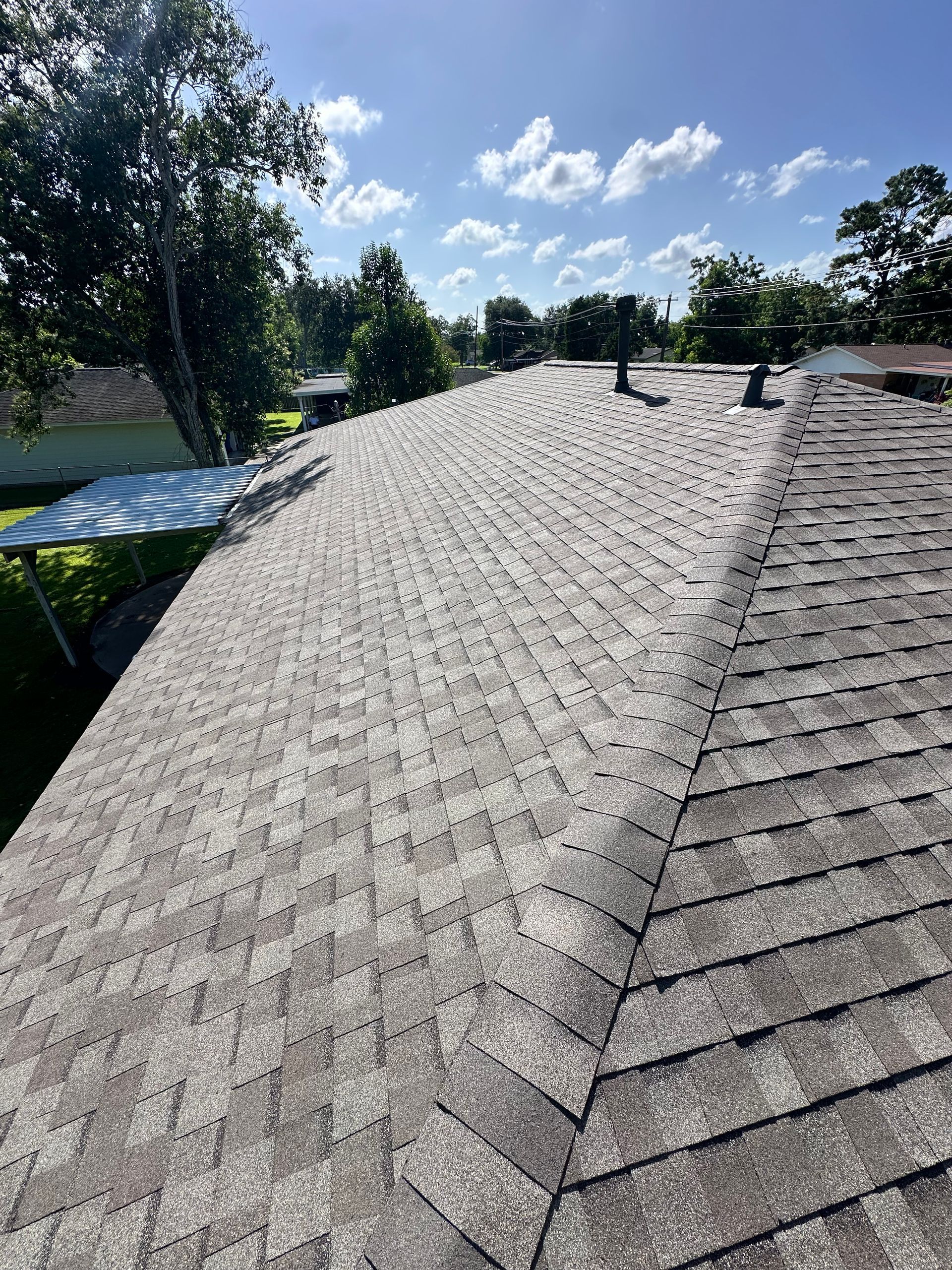 View of a gray shingled roof under a partly cloudy blue sky. Trees and a yard are in the background.