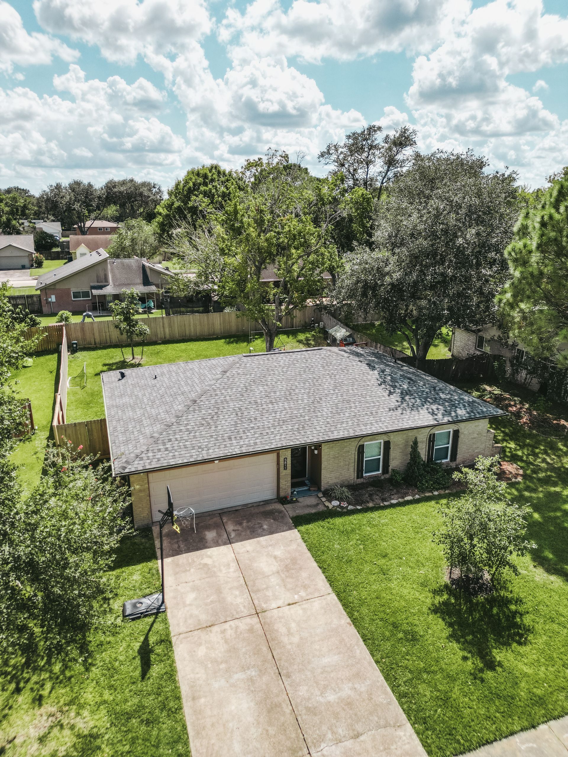 Aerial view of a single-story house with a driveway, green lawn, and trees under a partly cloudy sky