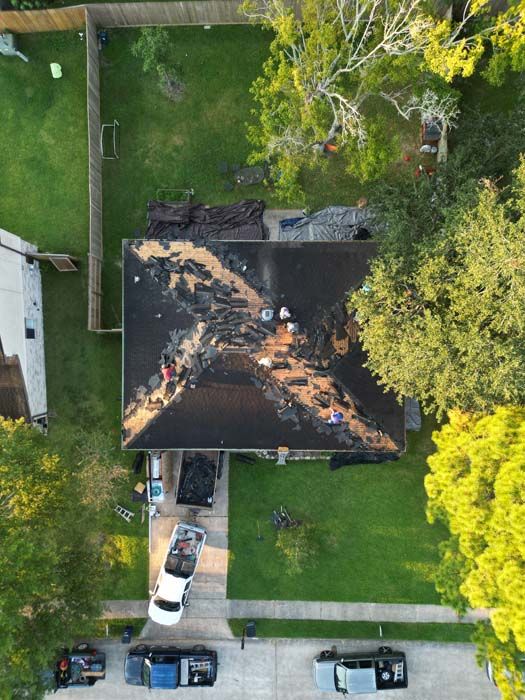Aerial view of a house with a dark roof, surrounded by green yard and parked cars along the street.