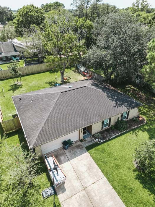 Aerial view of a single-story house with a driveway, green lawn, and large trees.