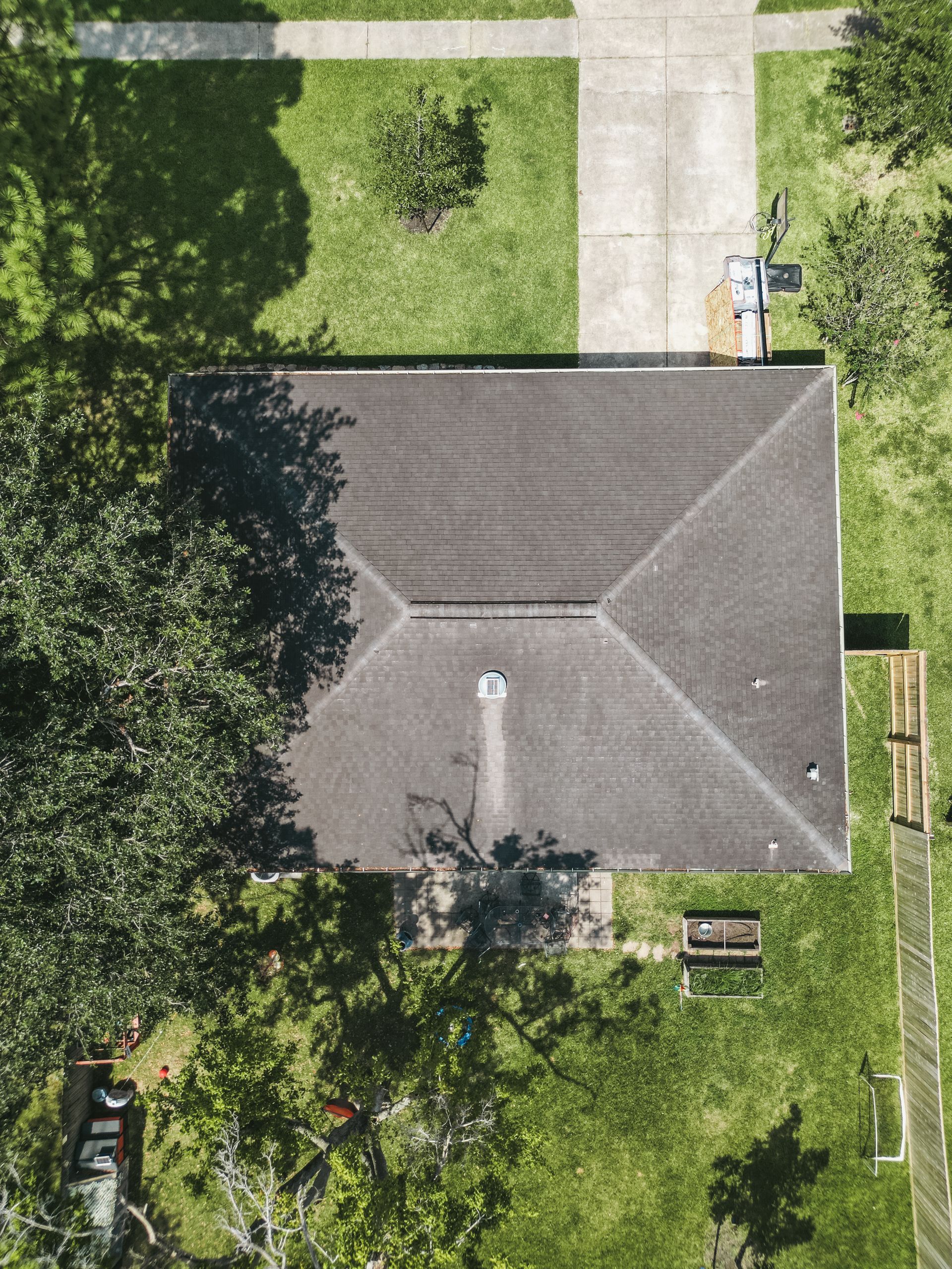 Aerial view of a gray-roofed house surrounded by green trees and lawn with a driveway and sidewalk.