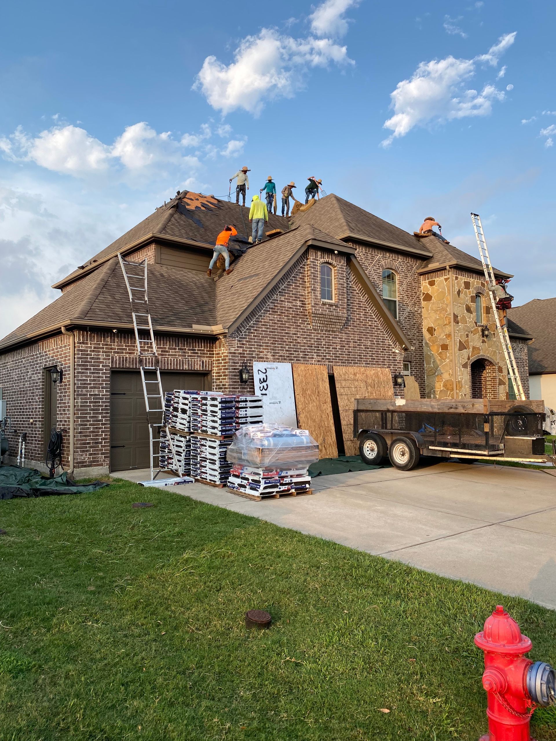 Roofers working on a house with pallets of shingles on the ground. Ladders and a trailer are nearby.