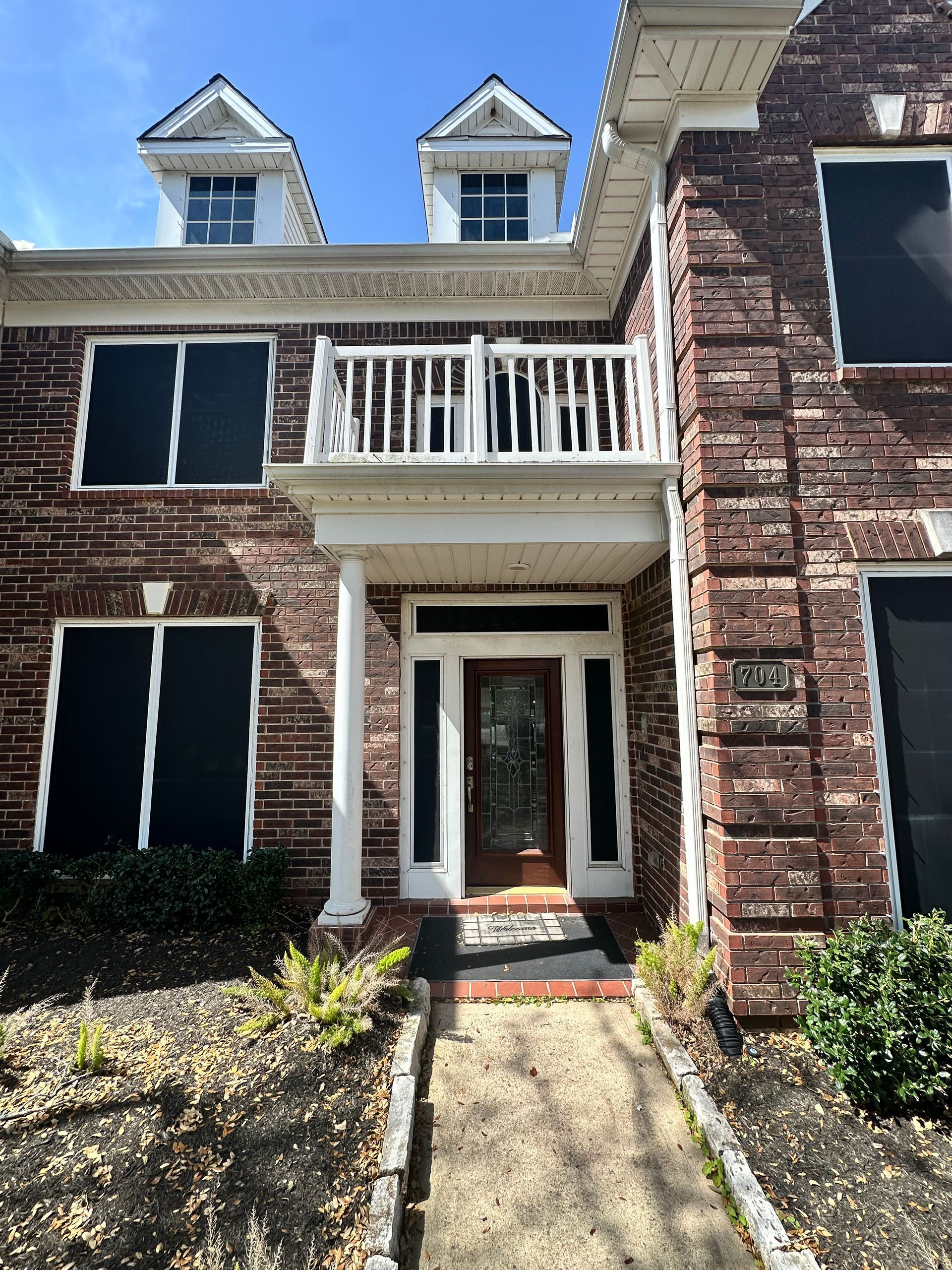 Brick house with black window coverings, balcony, and dormers under a blue sky.