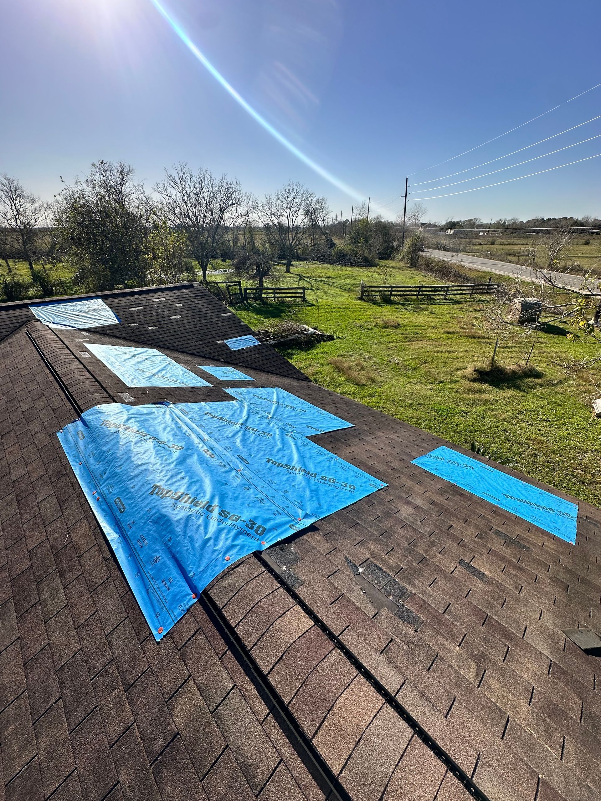 Blue tarps covering a damaged shingle roof under a bright sky, with a grassy field and trees in the background.