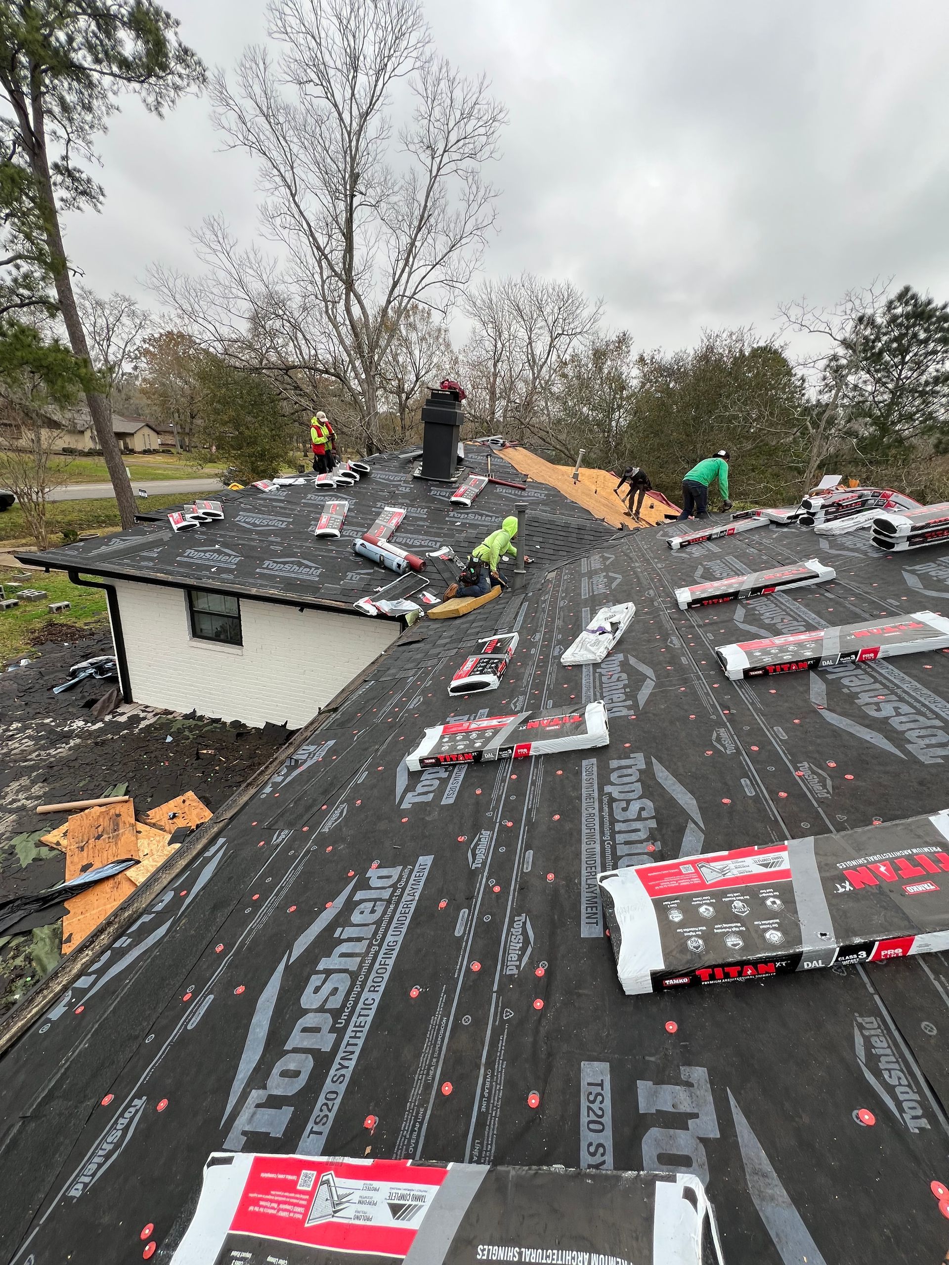Roofers working on a house roof, laying asphalt shingles and materials. Cloudy day, surrounded by trees.