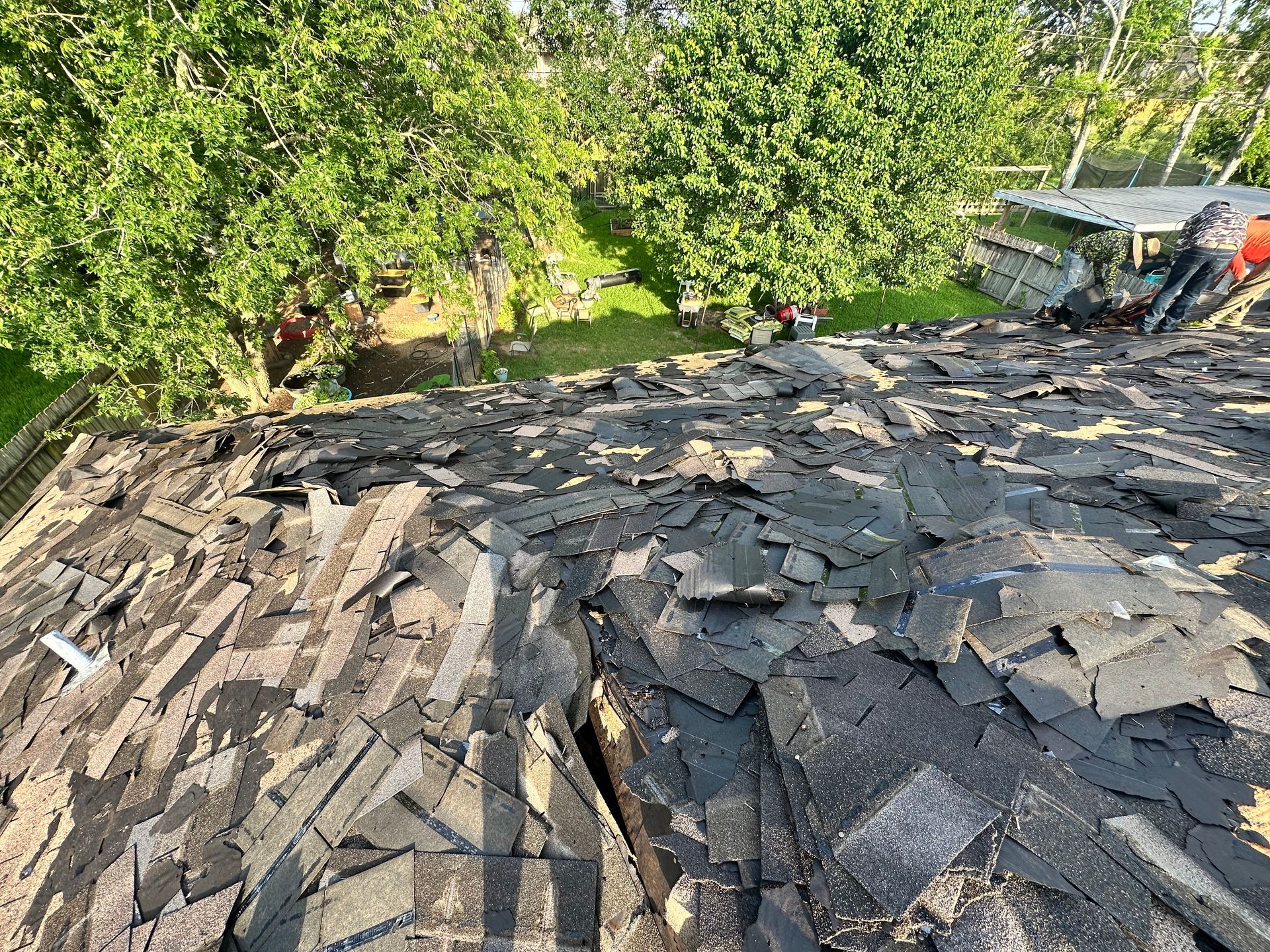 Damaged rooftop covered in torn shingles, with trees in background.