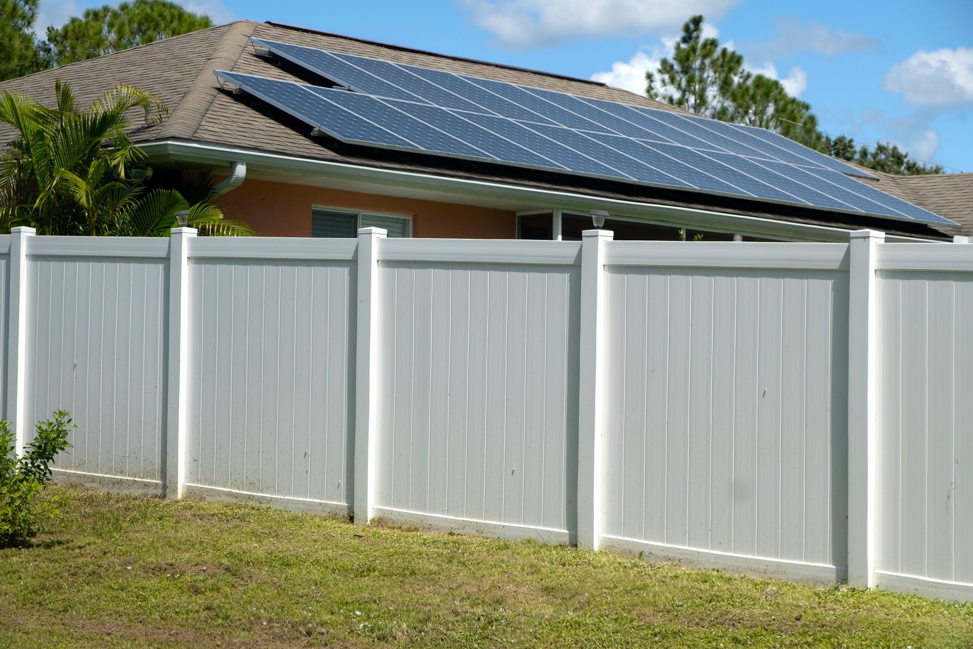 White fence in front of a house with solar panels on the roof under a partly cloudy sky.