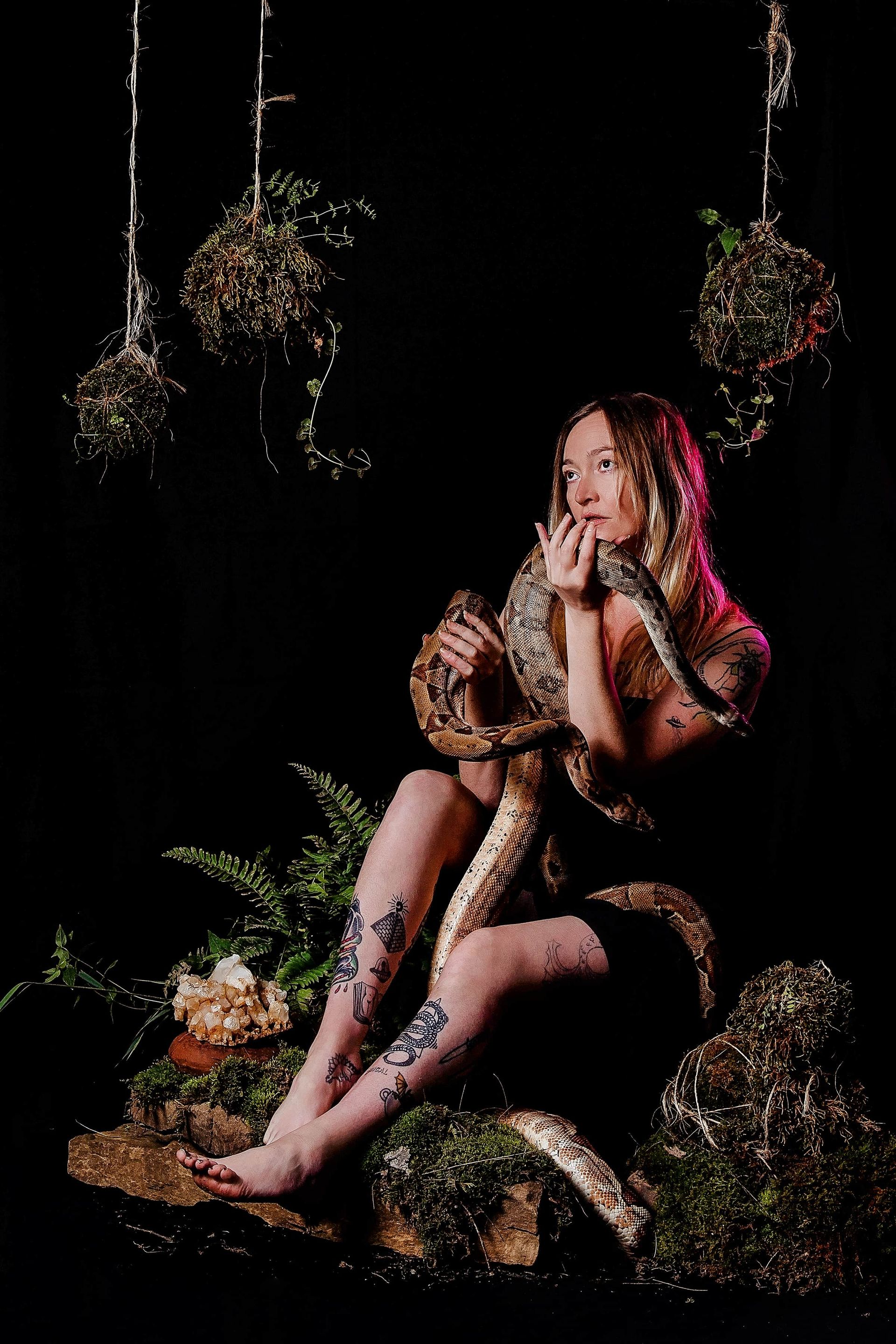 A woman is sitting on a rock with a snake around her neck.