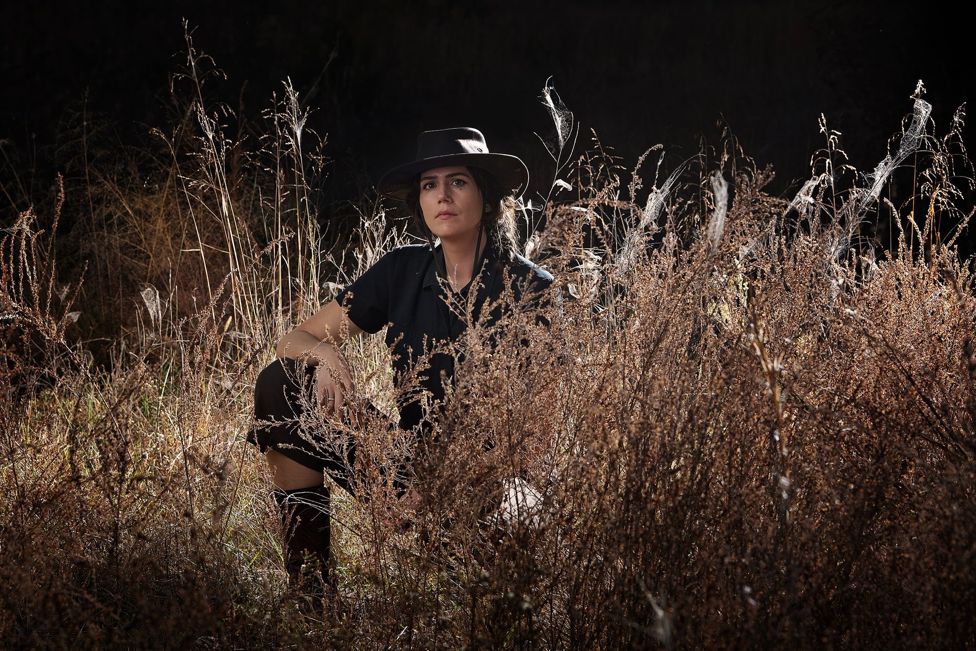 A woman in a black hat is kneeling in a field of tall grass.
