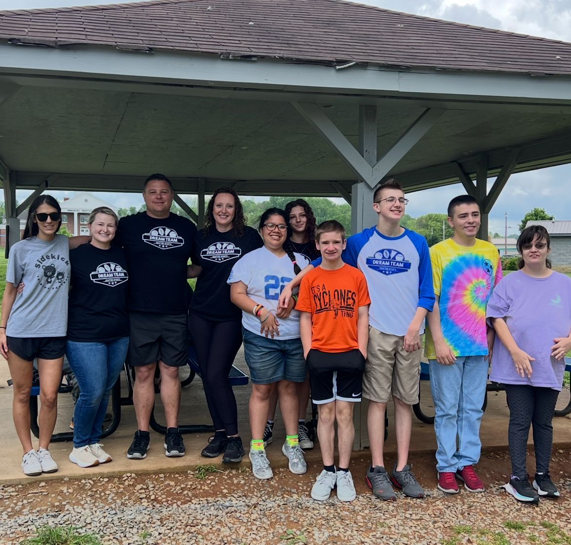 Group of people standing under a pavilion. They are wearing t-shirts. Cloudy sky.