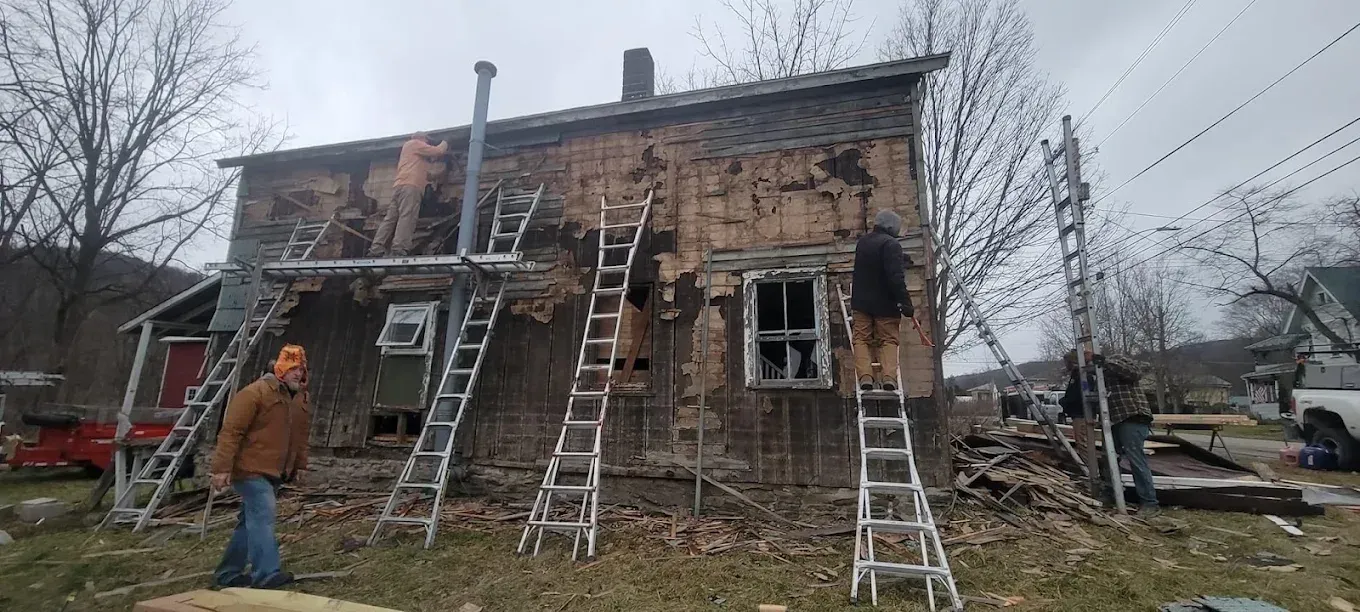 Worker on a ladder repairing a charred two-story house exterior with multiple ladders set up.