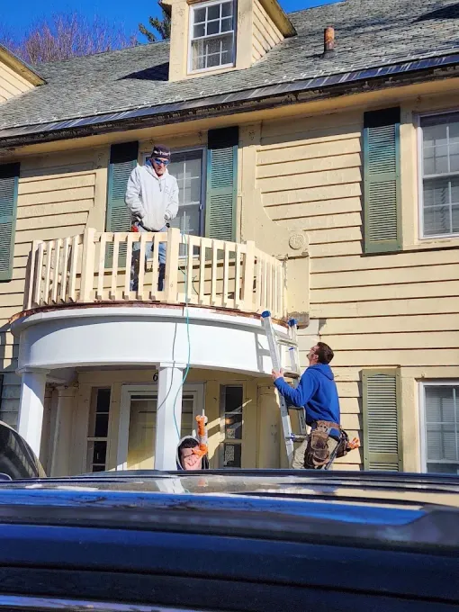 Two workers repairing a beige house balcony and front porch, one on the balcony and one on a ladder.