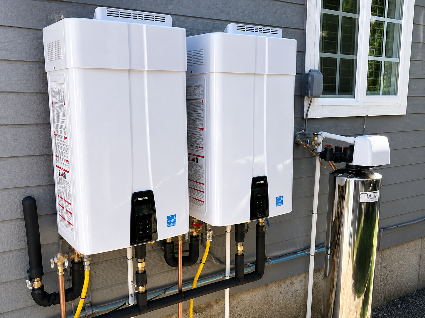 Two white wall-mounted tankless water heaters with pipes and vents outside a house
