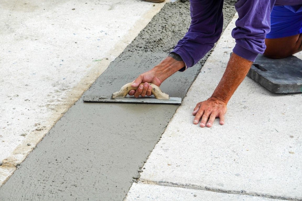 Person using a trowel to smooth wet cement in a narrow trench between concrete slabs.