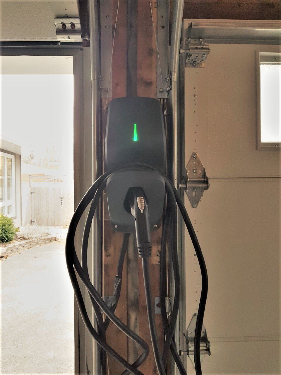 Borne de recharge pour véhicule électrique fixée sur un mur en bois d'un garage, avec un voyant vert allumé et un câble qui pend.