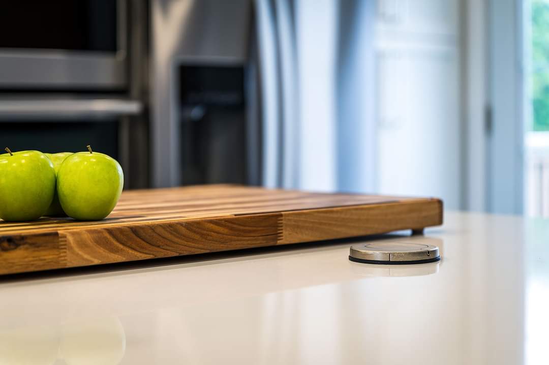 Gros plan sur un plan de travail blanc avec une planche à découper en bois et des pommes vertes. Un petit objet métallique se trouve au premier plan.
