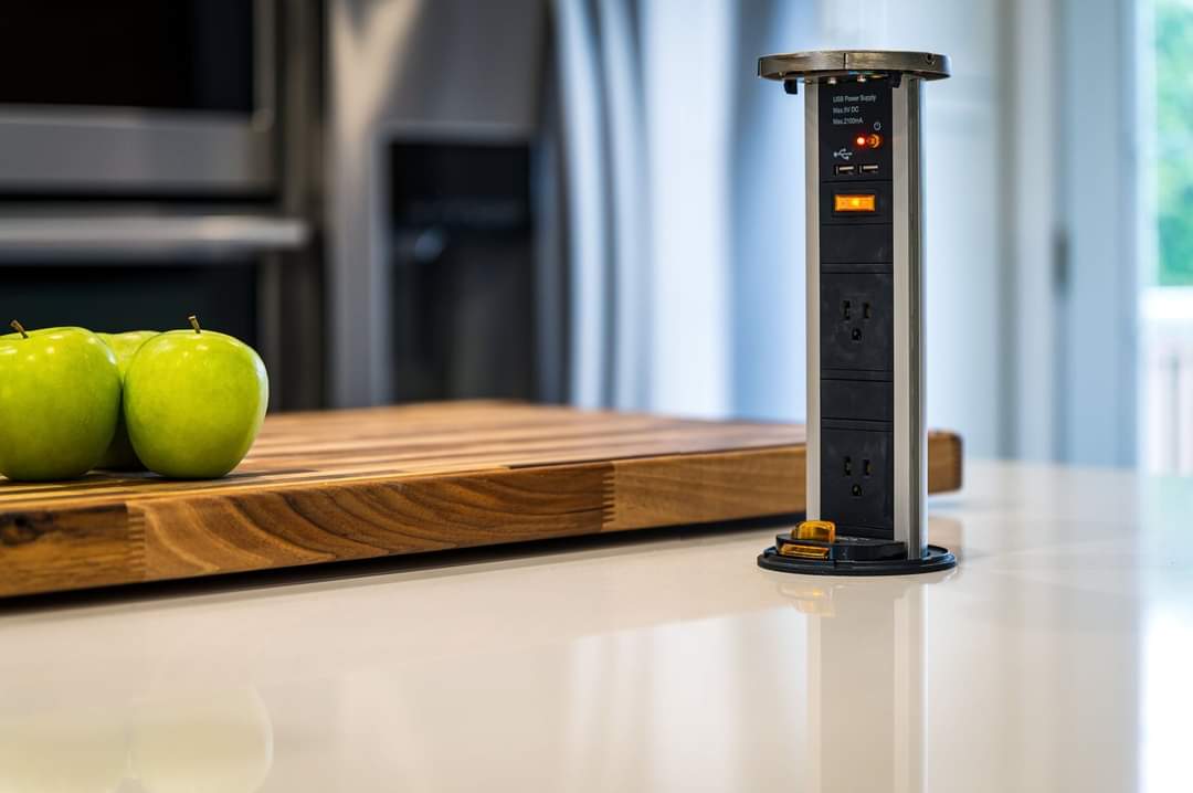Plan de travail de cuisine avec une prise électrique escamotable à côté d'une planche à découper en bois avec des pommes vertes.