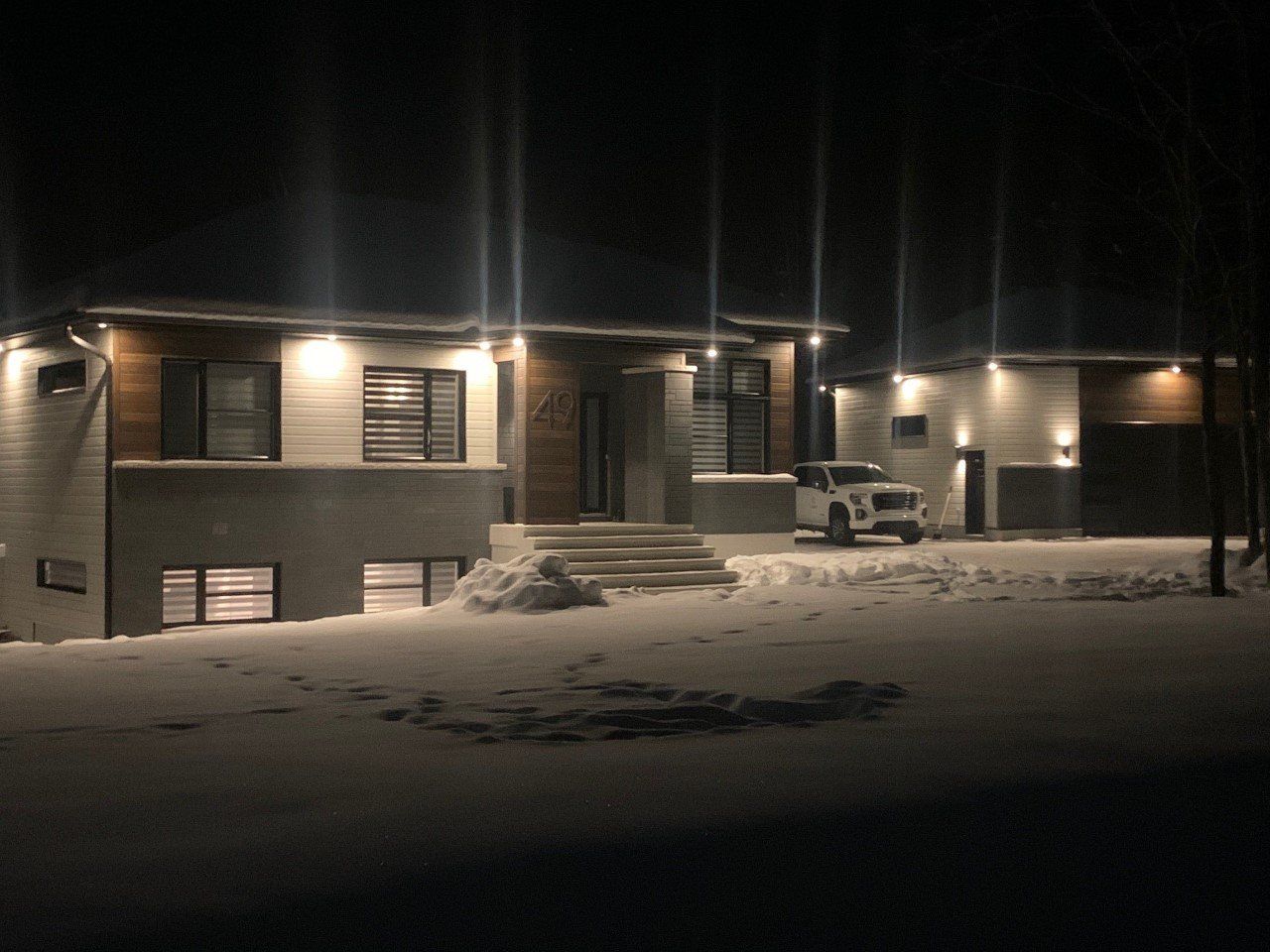 Maison moderne la nuit, extérieur éclairé, sol enneigé.