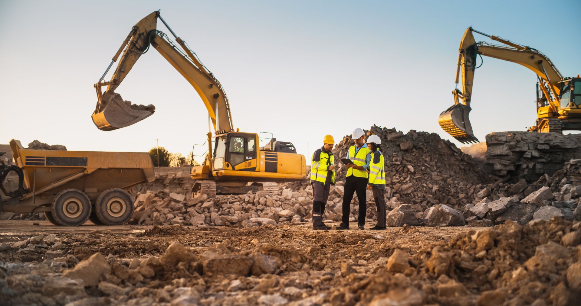 Construction site with excavators and workers in yellow safety vests on a dirt mound at sunset
