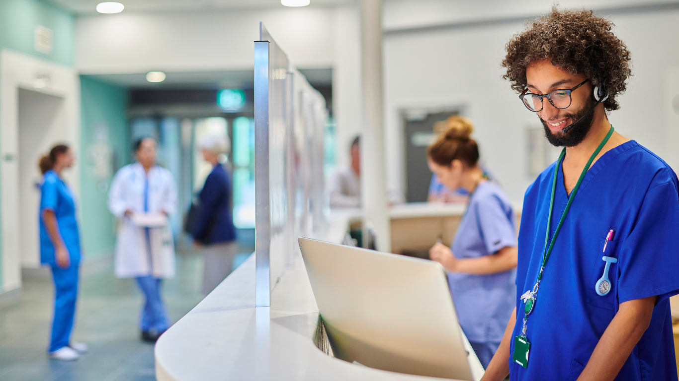 Healthcare worker in blue scrubs reviewing a chart in a hospital corridor with staff in the background