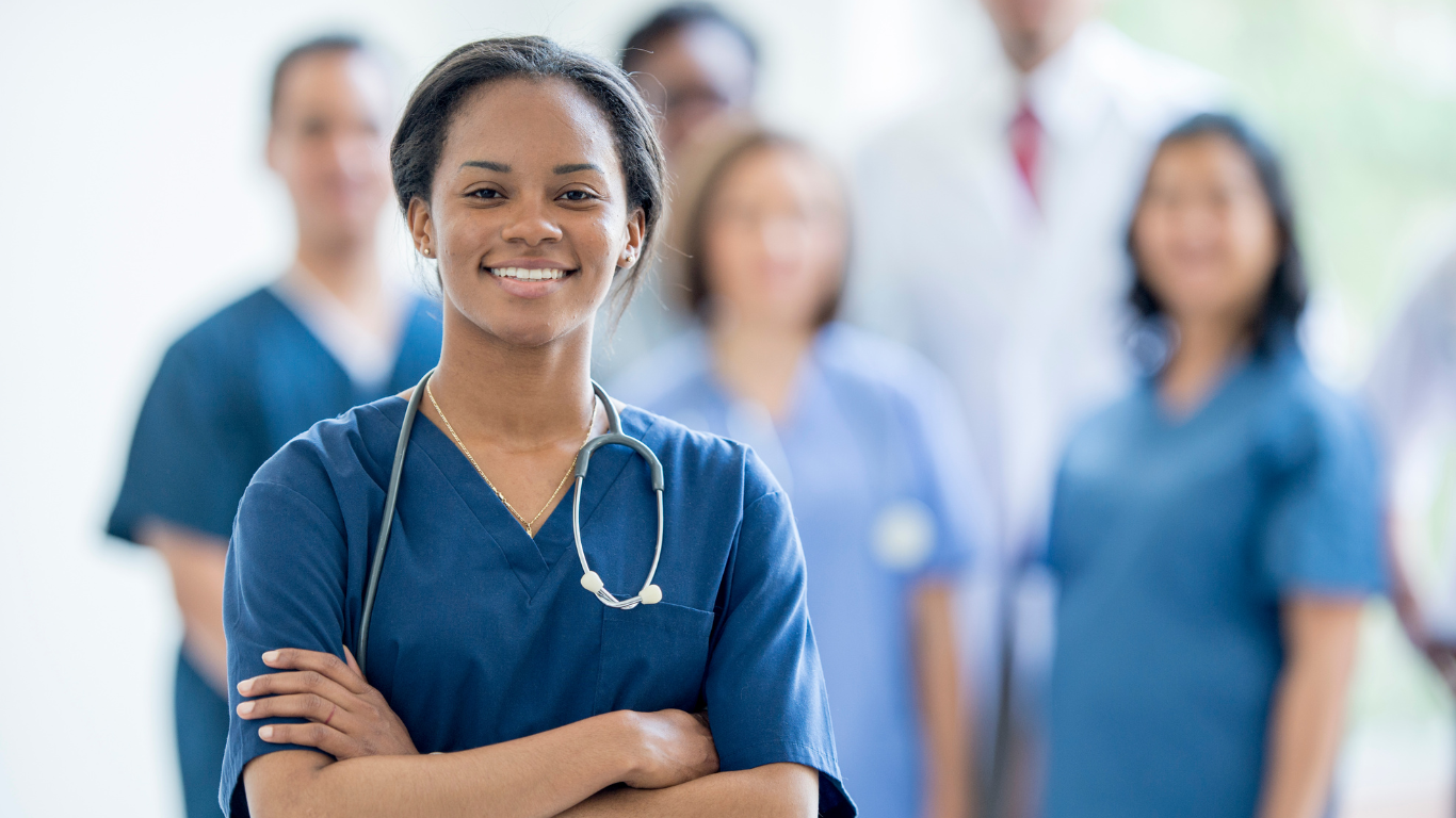Smiling healthcare worker in blue scrubs with stethoscope, standing in front of blurred medical staff.