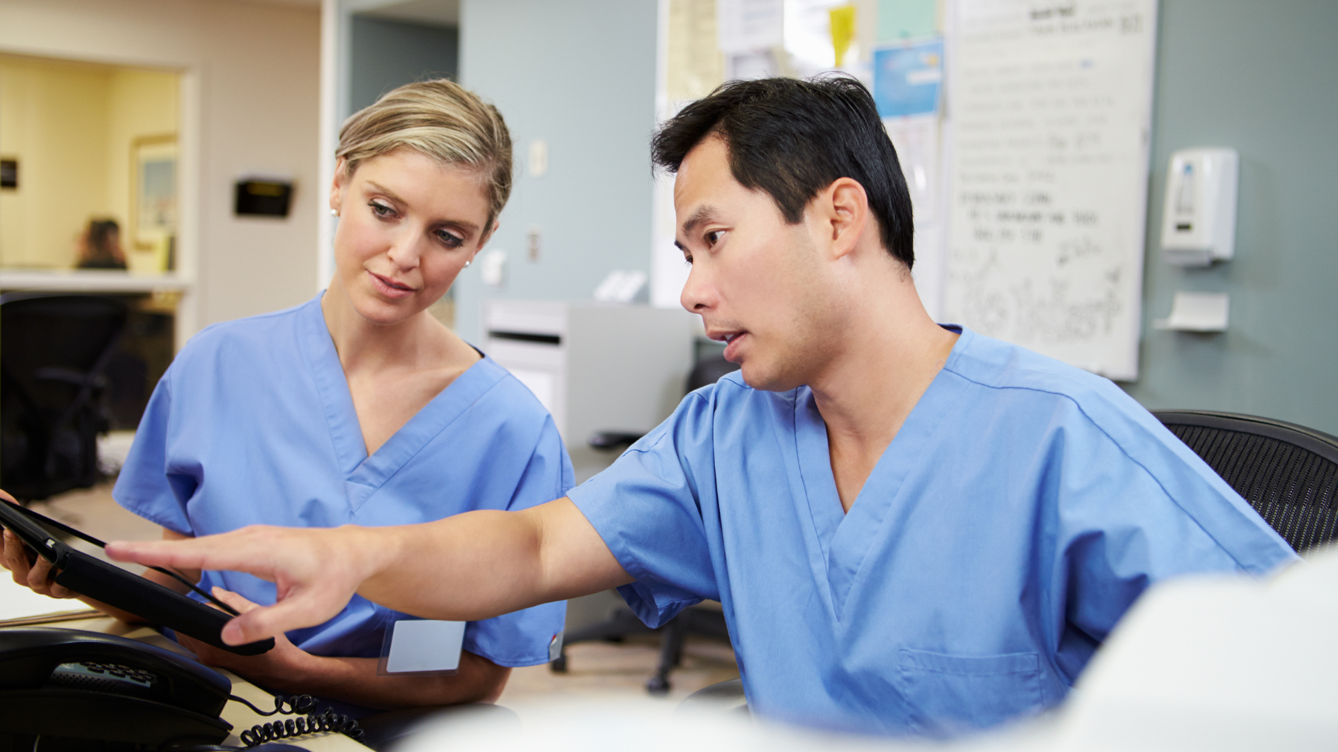 Two healthcare workers in blue scrubs reviewing a tablet together in a clinic office