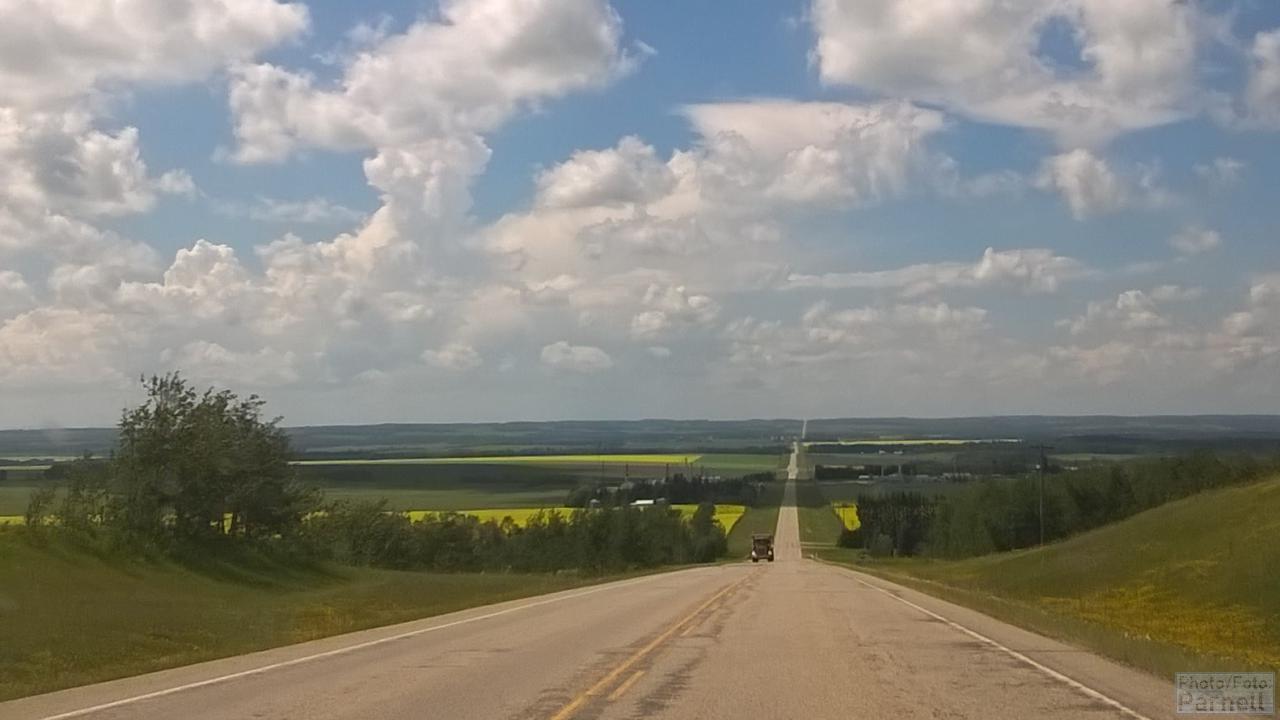 View across a wide valley with alternating farm fields of grain and canola (rapeseed) and several farmyards.