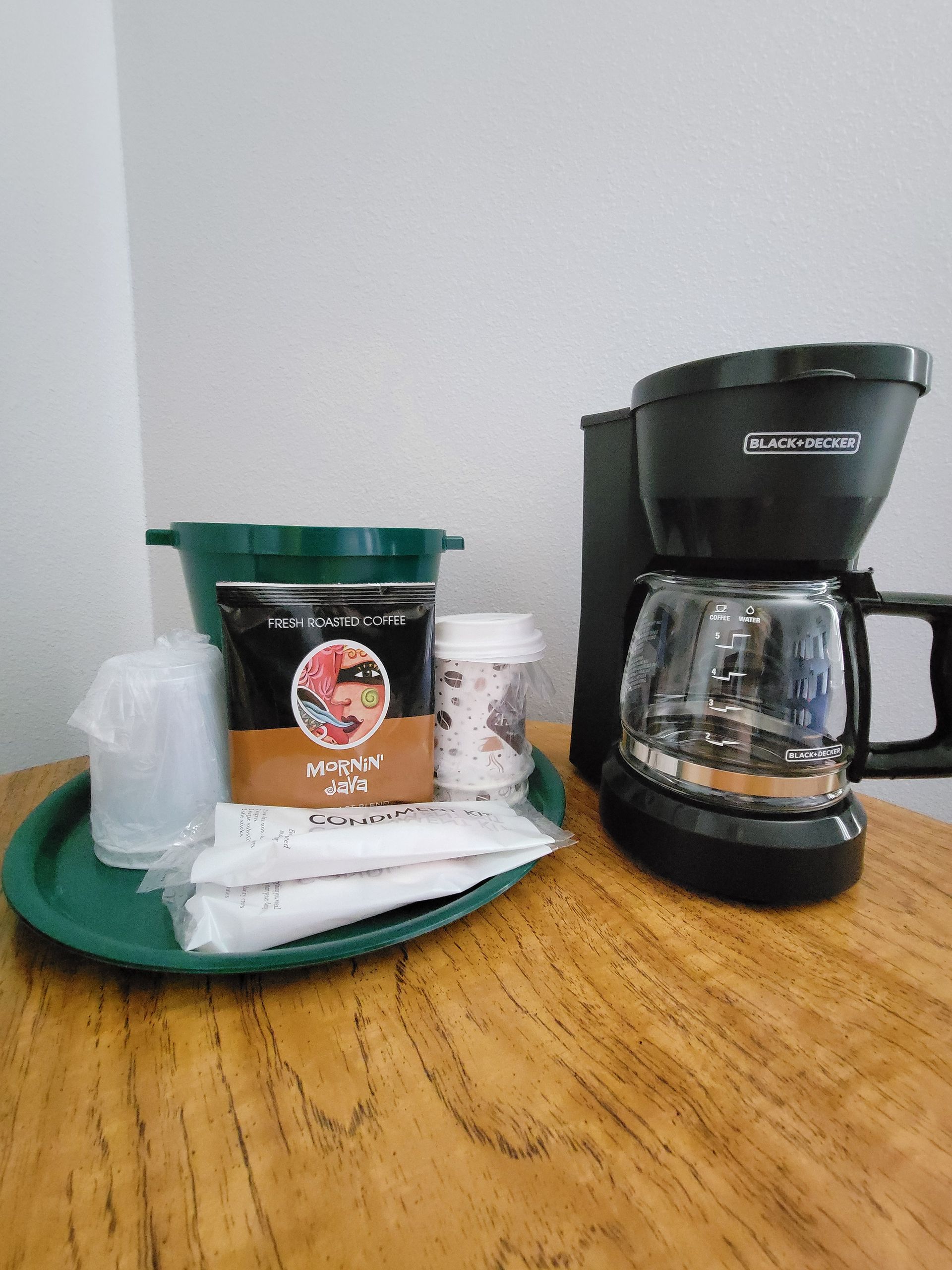 Coffee maker and supplies on a wooden table, including a coffee pot, coffee mix, cups, and a green tray.
