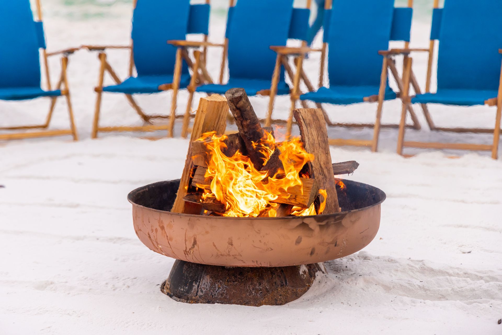 Fire pit with burning wood on a sandy beach, blue chairs in background.
