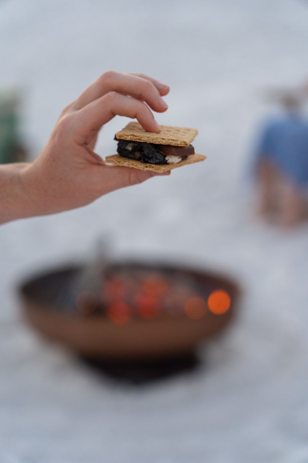 Hand holding a s'more above a fire pit with lit embers.