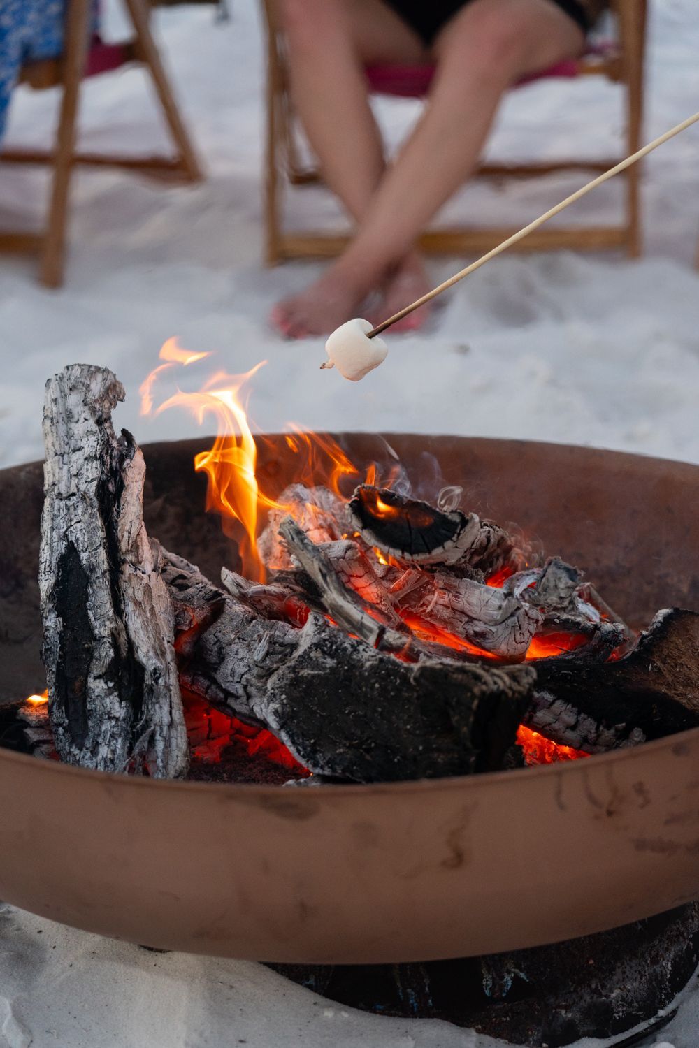 Marshmallow roasting over a fire pit on a beach. A person's legs and chairs are visible in the background.