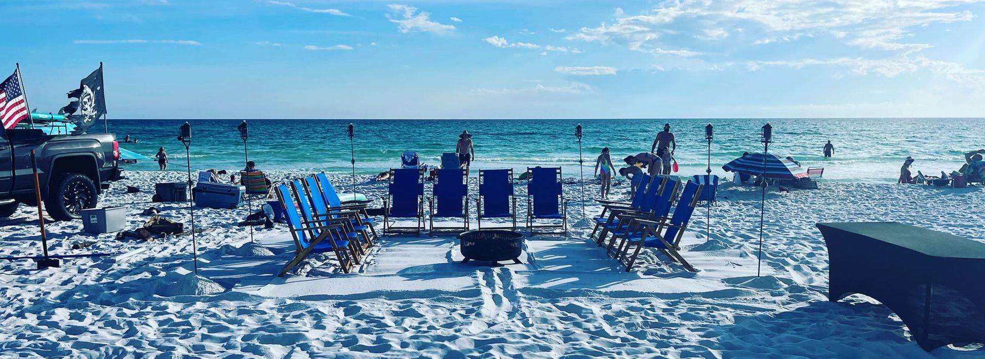 Beach scene with blue chairs, fire pit, and people enjoying the water and sun. American and skull flags on a truck.