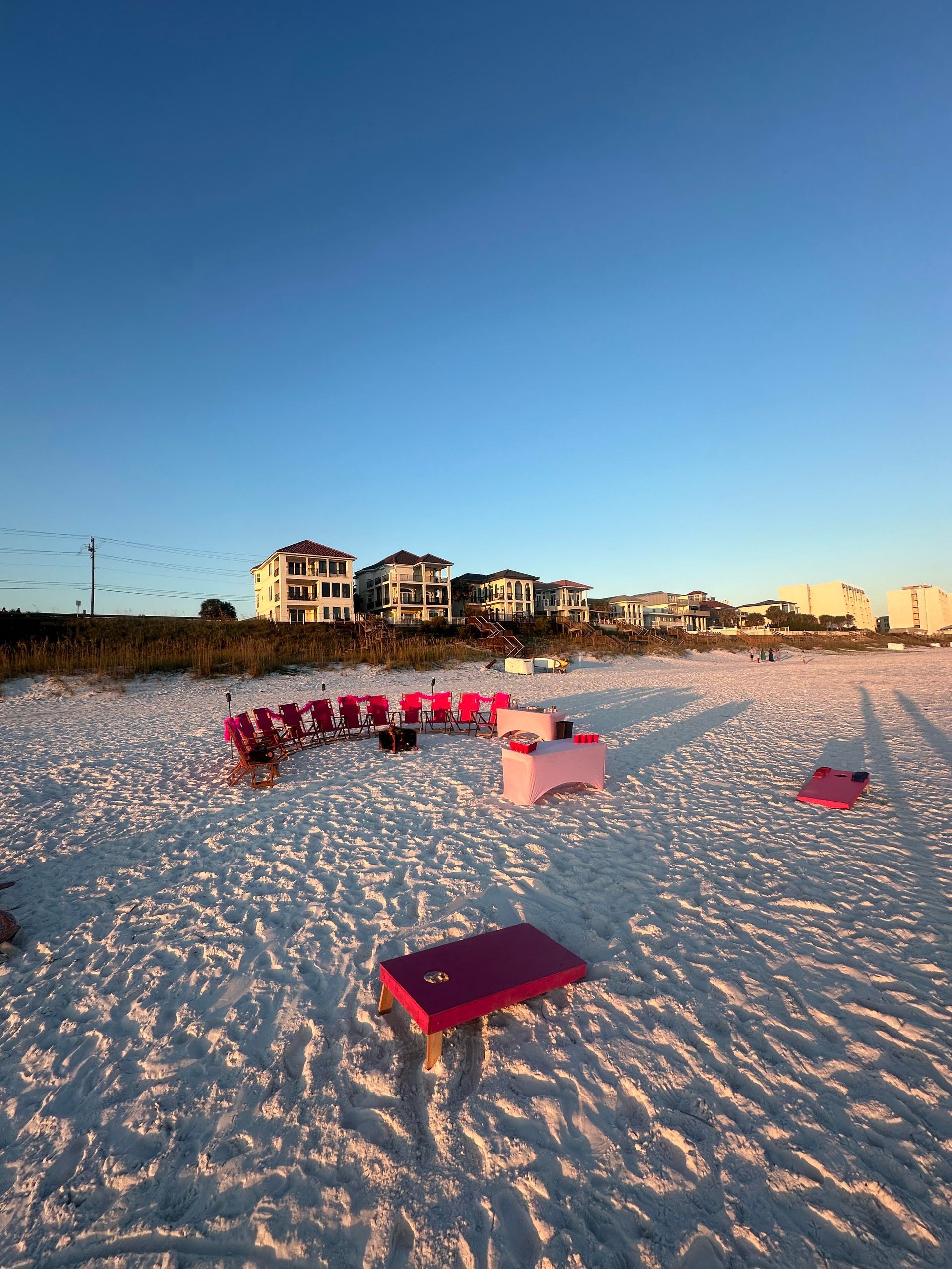 Beach scene with pink chairs and tables on white sand, houses in background under blue sky.