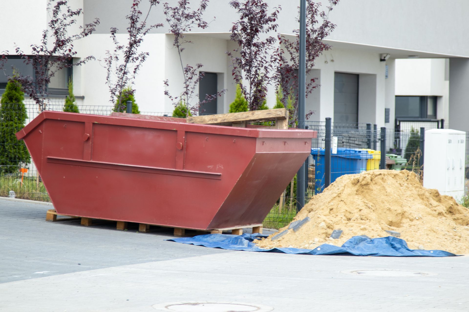 A dumpster showcasing efficient dumpster rental filled with construction rubble at job site. Red dumpster on a pallet, beside a pile of sand, in front of a white building.