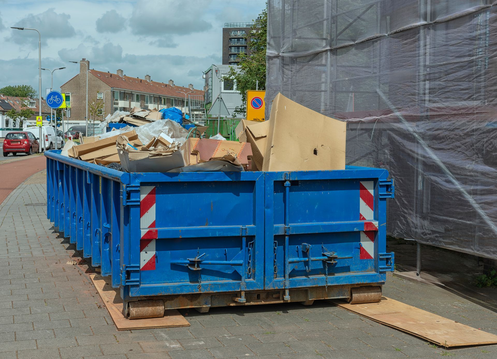 Blue dumpster filled with debris on a city sidewalk, next to a building covered in plastic sheeting.