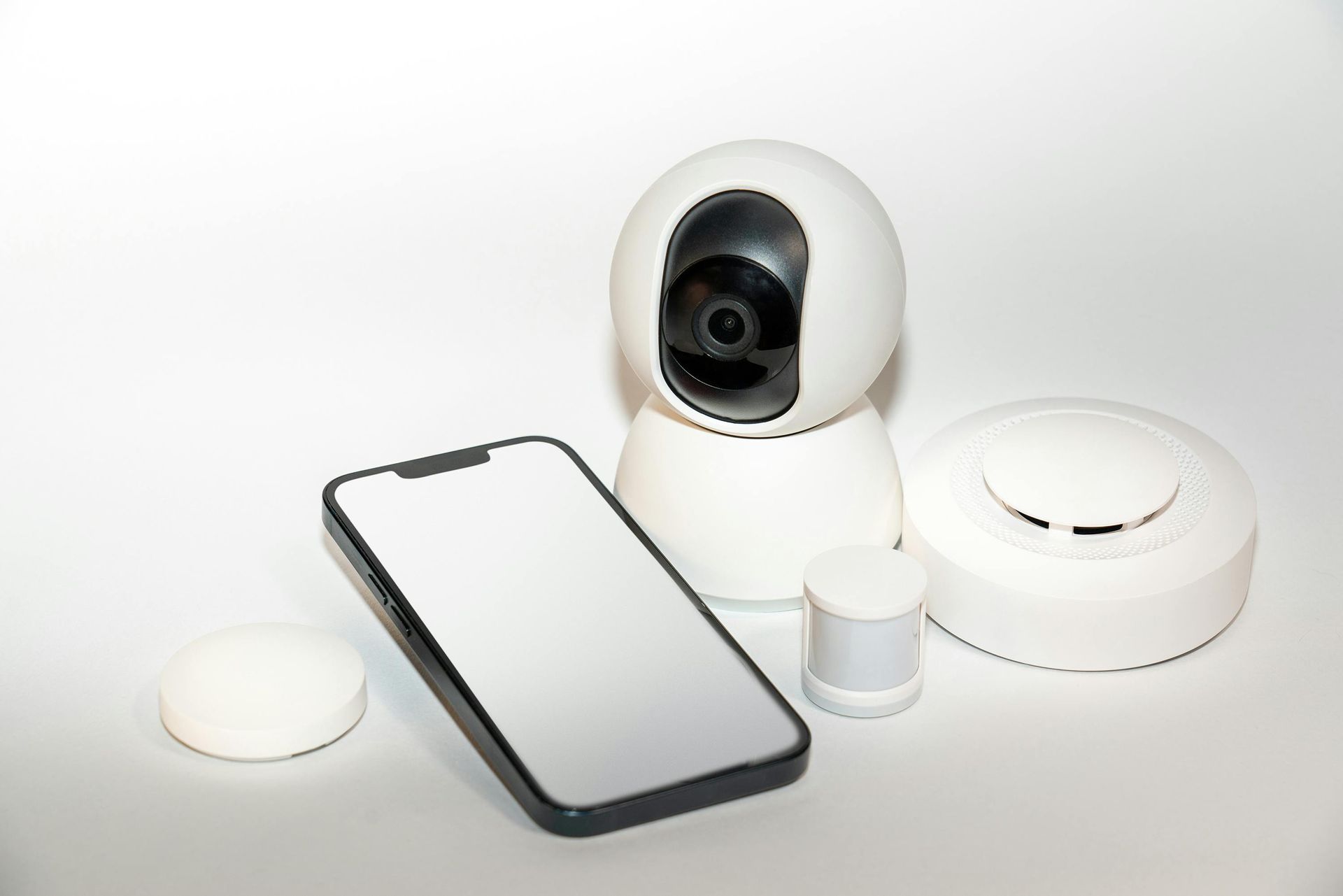 A white smart security camera, smartphone, motion sensor, and hub arranged on a plain white surface.
