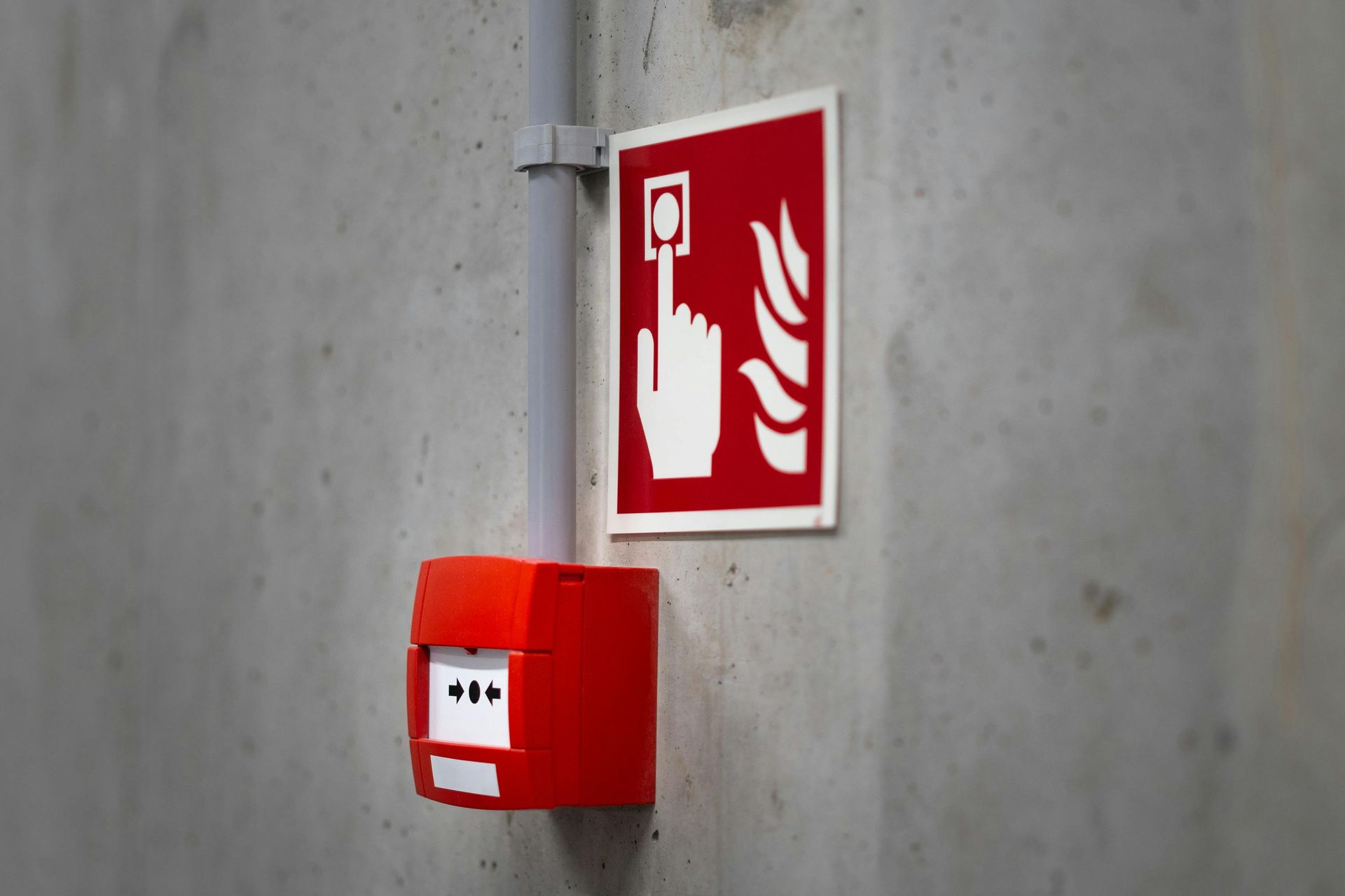 A red fire alarm manual call point mounted on a concrete wall, accompanied by a sign showing a hand pressing a button.