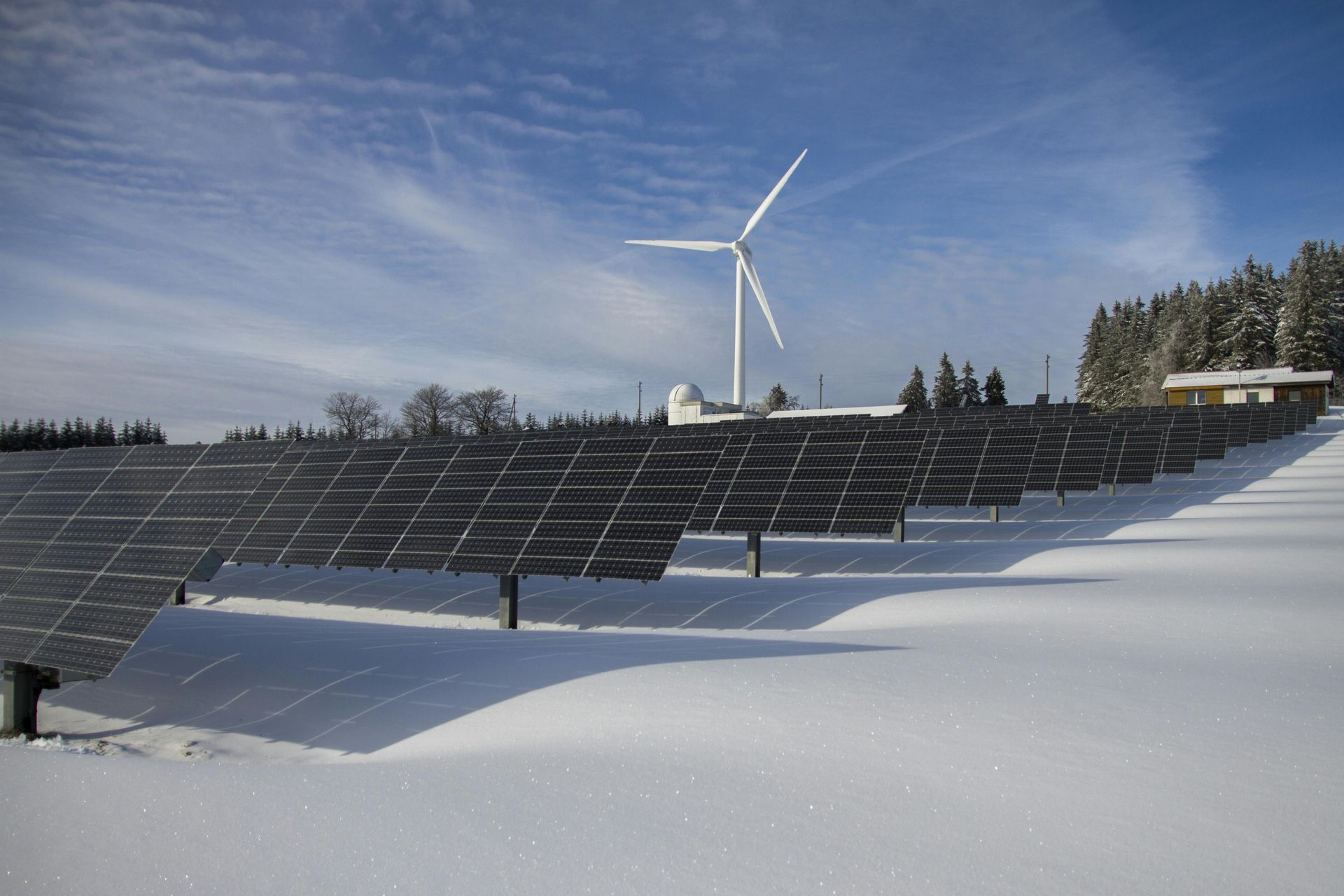 A wind turbine and rows of solar panels sit in a snow-covered field under a bright blue sky.