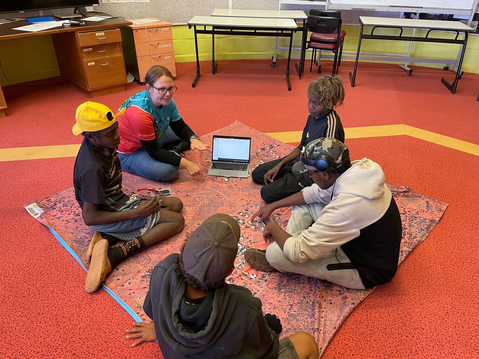Students sit in a circle on a rug around a laptop in a classroom.