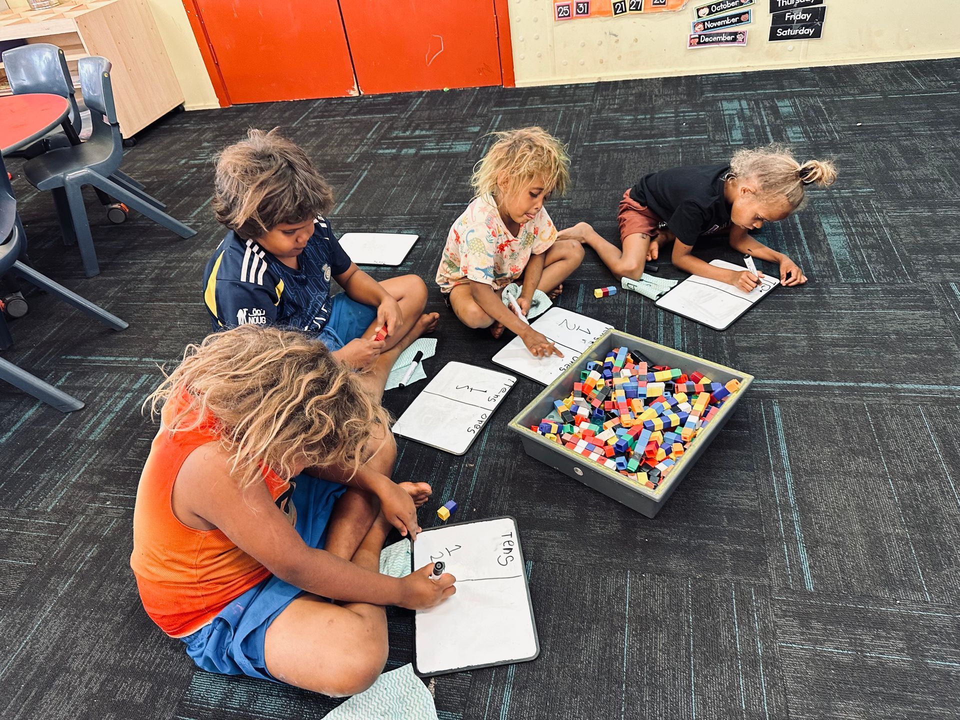 Children drawing on whiteboards on a classroom floor beside a tray of colorful blocks.