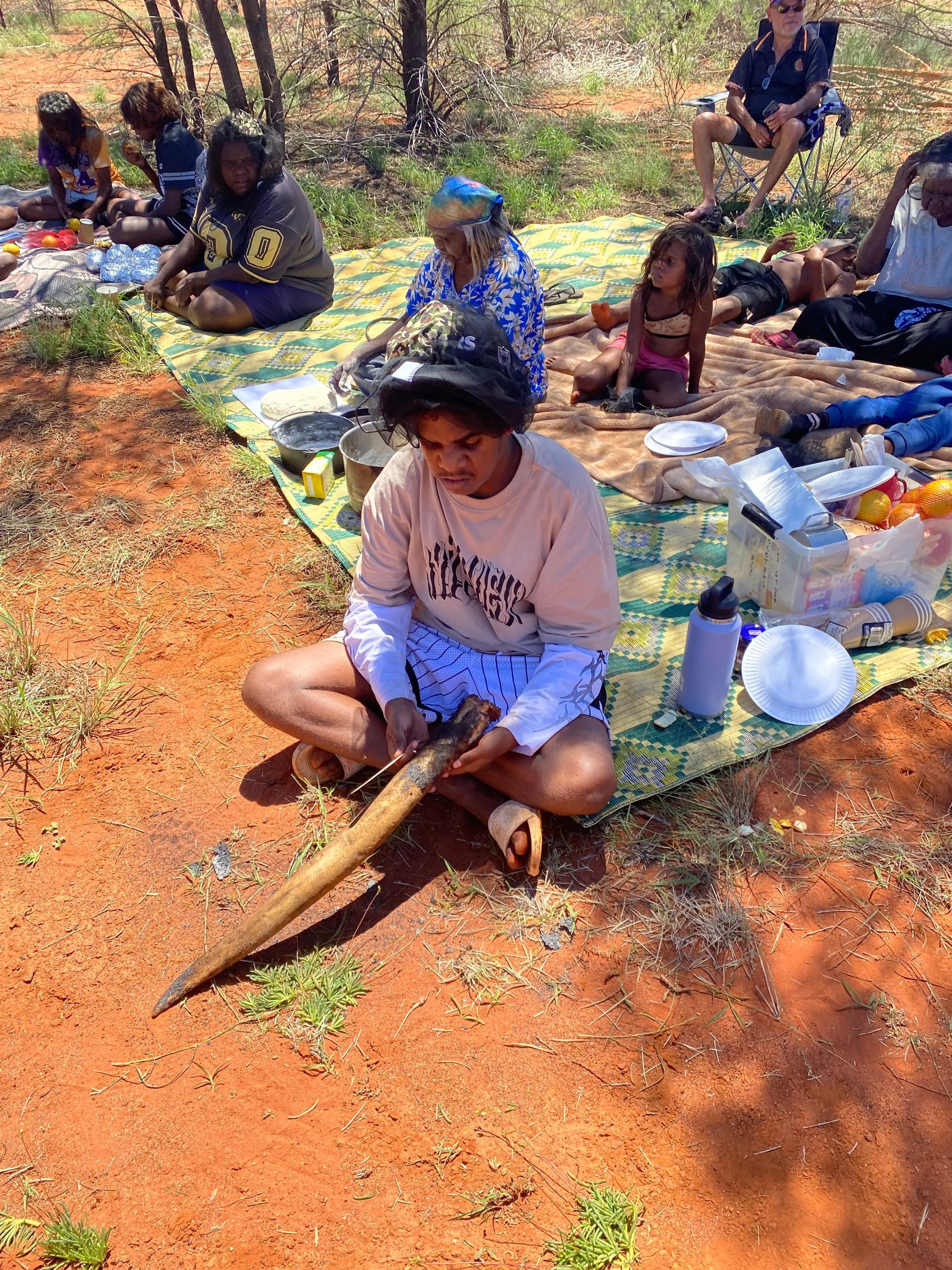 People seated outdoors on mats, making or sorting materials on red dirt.